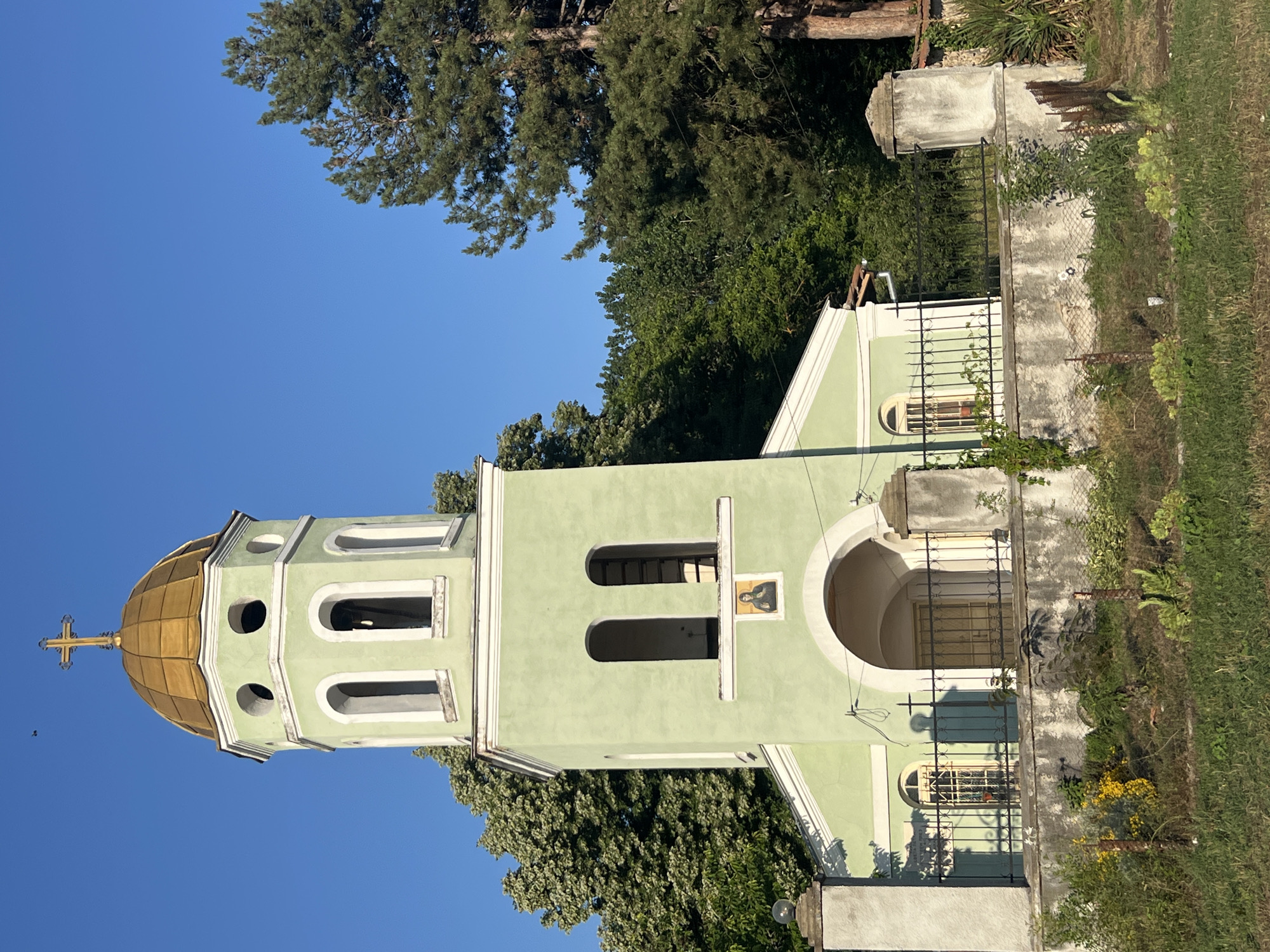 The image shows a pale green church with a golden dome, topped with a cross. The church has arched windows and an arched entrance. The scene is set outdoors with a clear blue sky visible above. Lush green trees surround the church, and there is a grassy area in the foreground with some plants and a chain-link fence. A concrete pillar with a weathered texture stands to the right of the church, and a smaller concrete structure is visible in the lower foreground. The lighting suggests it is daytime, possibly morning or late afternoon, with long shadows cast by the church. There are no people or animals visible in the image. The style of the church, particularly the dome and cross, suggests it is an Orthodox Christian church. The overall impression is one of a serene and somewhat rustic religious building in a natural setting.