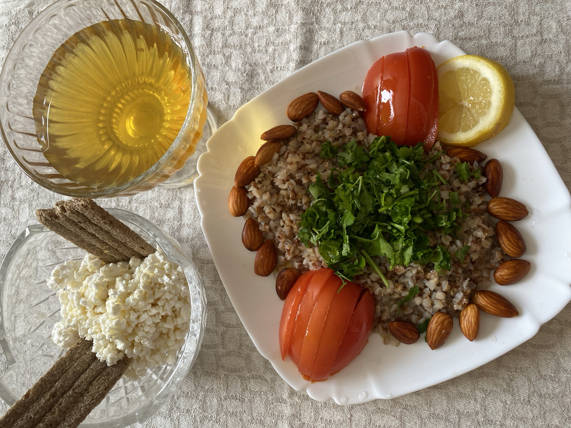 The image shows a table laid with a meal. In the foreground, a white plate holds a portion of cooked buckwheat, garnished with fresh parsley. Several almonds are scattered around the buckwheat. Two slices of tomato, one on each side of the buckwheat, and a slice of lemon are also present on the plate. In the background, a glass bowl contains cottage cheese with two pieces of dark bread. Another glass bowl holds a golden liquid, likely honey. The scene is set on a textured, beige tablecloth. There are no people visible in the image. The image does not provide any information about the location in the city of Lida, Belarus.