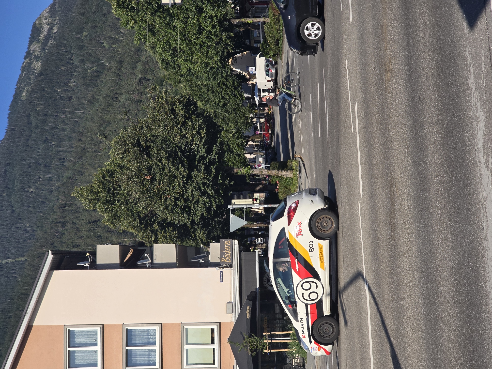 In Garmisch-Partenkirchen, Germany, a white Ford Ka, adorned with racing decals and the number 69, is observed traveling on a paved road. The car displays "Würth" branding on its side, along with "Twix" and other logos. Inside the vehicle, a man with glasses is visible in the driver's seat. In the background, a bakery with a sign reading "Bäckerei" is situated next to a multi-story building. Further in the distance, a forested mountain rises under a clear blue sky. A man on a bicycle rides across the street, while other cars are present on the road. The scene depicts a street-level view of a town, with elements of both urban and natural scenery.