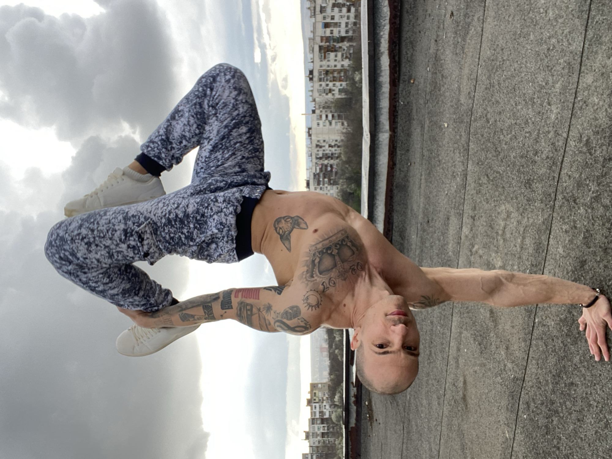 A bald man with tattoos is performing a handstand on a rooftop in Pleven, Bulgaria. The man is wearing sweatpants and white sneakers. He is shirtless, revealing several tattoos on his chest and arms. The rooftop is made of gray material and has lines running across it. In the background, there are buildings and a cloudy sky. The man is looking directly at the camera.