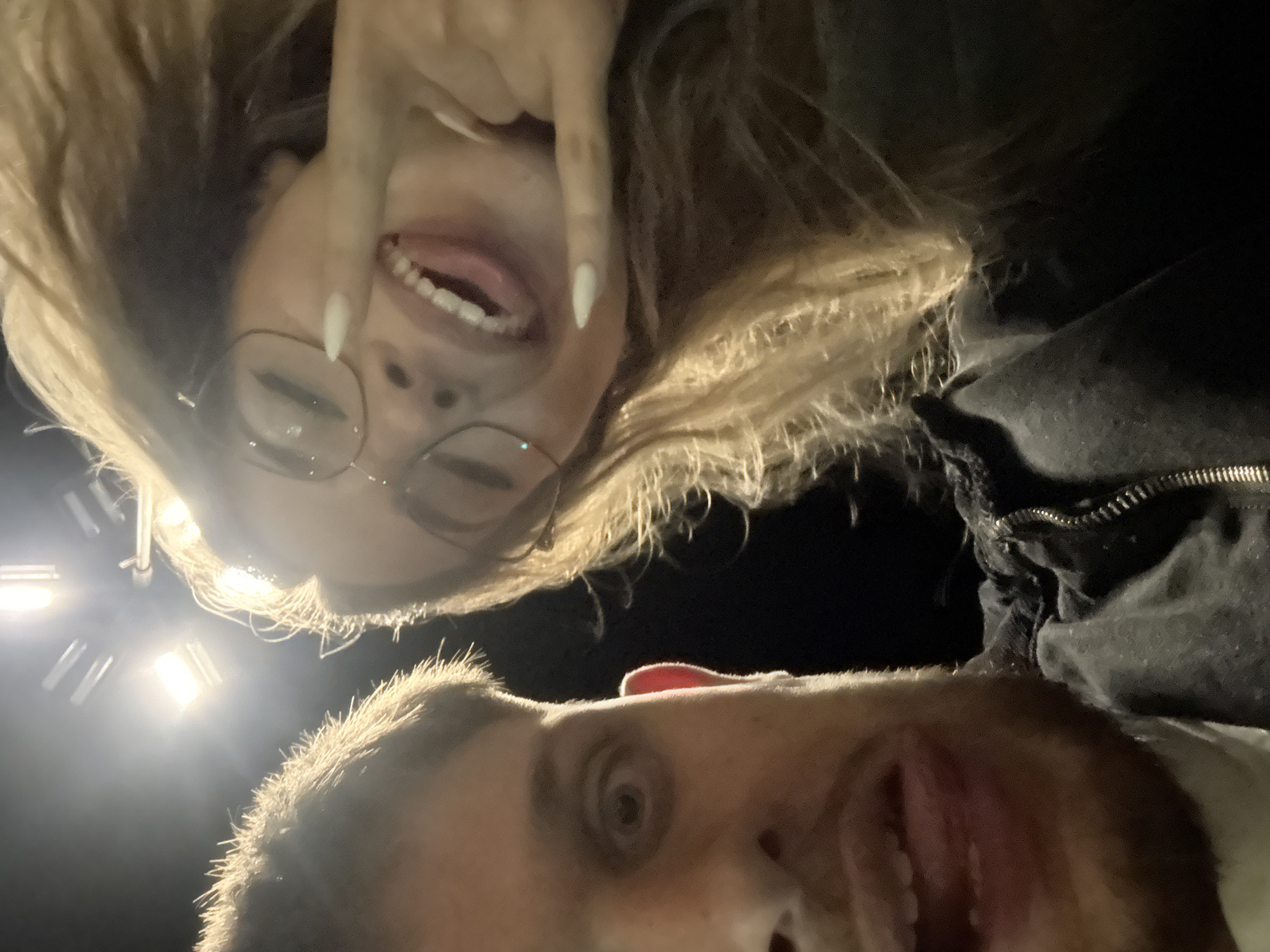 Two people are taking a selfie under bright lights. The person on top, appearing female, has long blond hair, wears glasses, sticks out their tongue, and holds up two fingers in a peace sign. The person on the bottom, appearing male, has short brown hair, a beard, and also sticks out their tongue. The location is likely at night, based on the dark background. There is no indication it is in Blagoevgrad, Bulgaria.