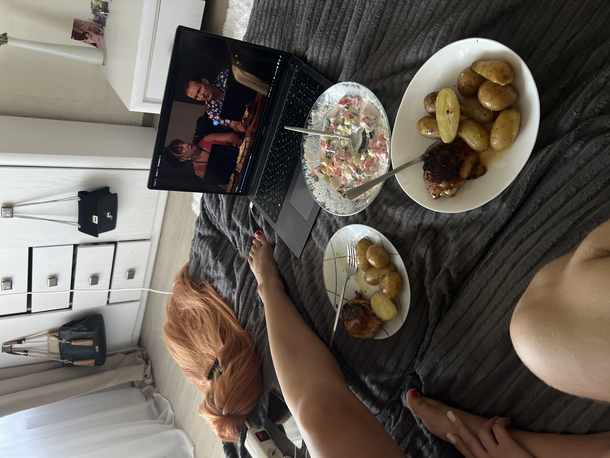 The image shows a person relaxing on a bed, watching a movie on a laptop. The bed is covered with a dark gray, textured blanket. Next to the laptop are two plates of food. One plate contains several small, cooked potatoes and a piece of grilled chicken. The other plate holds a salad made with chopped tomatoes, cucumbers, and a creamy dressing, with a spoon in it.
The laptop screen displays a scene from a movie or TV show, featuring two people. One person is wearing a patterned shirt, and the other is wearing a pink top. The scene appears to be set indoors.
In the background, to the left, there is a white dresser with several drawers. A black handbag with a silver chain strap is placed on top of the dresser, and another similar handbag is on the bottom drawer. A white vase with a plant is also on top of the dresser, along with a few framed pictures. The room has curtains, suggesting it might be daytime, though the lighting is not very strong.
The person's legs are visible in the foreground, indicating they are lying down. One foot, with red nail polish, is positioned near the plate of food. A long, reddish-blonde wig is also visible on the bed. The overall scene suggests a relaxed, casual setting, likely at home. There are no visible weather cues or explicit time of day indicators beyond the presence of indoor lighting and curtains. No text is clearly legible in the image, other than what might be on the laptop screen, which is too small to read.