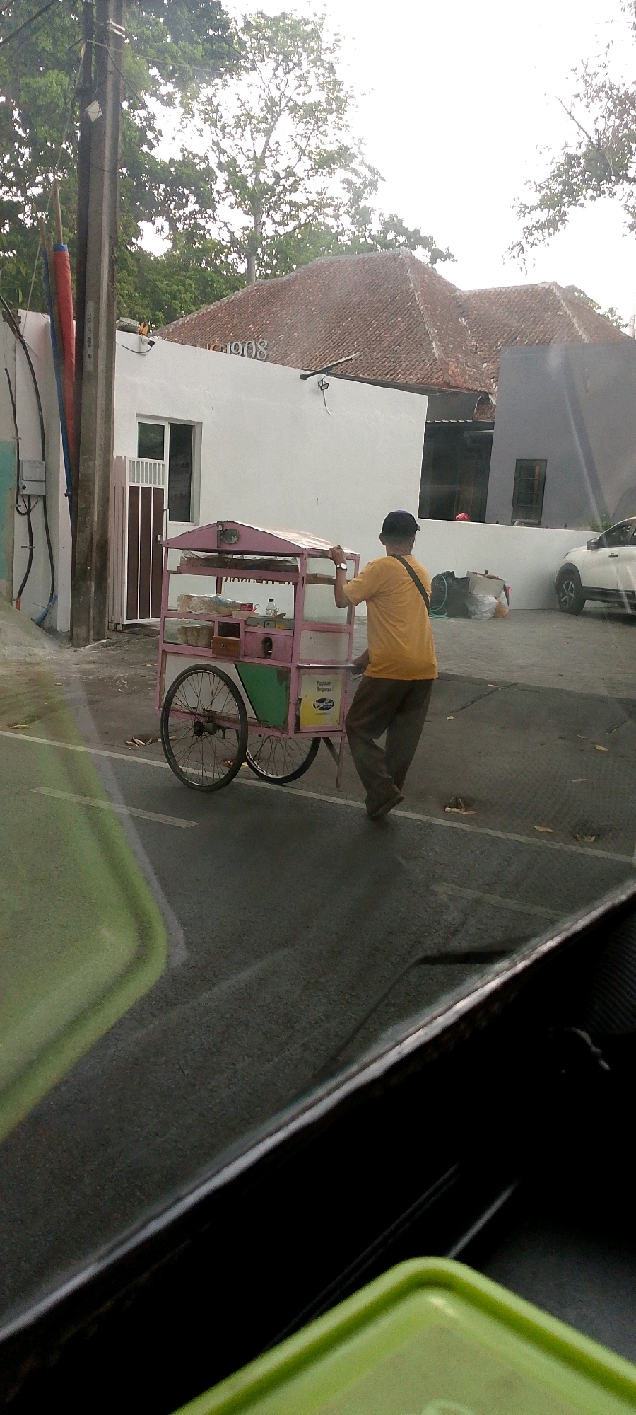 A man wearing a yellow t-shirt and dark pants pushes a pink and white food cart with two wheels. The cart has a covered top, shelves with items, and a green panel on the side. He is walking away from the viewer on a paved surface in front of a white building with a red tile roof. The building has windows and a door with vertical slats. A utility pole with wires is visible to the left of the building. Trees with green leaves are in the background. The scene appears to be outdoors in an urban or suburban setting. The image has a slightly blurry quality, possibly due to being taken from a moving vehicle. There is a hint of a green object in the foreground, likely part of the vehicle from which the photo was taken. The man's face is not visible. The building's roof has the number "1908" visible.