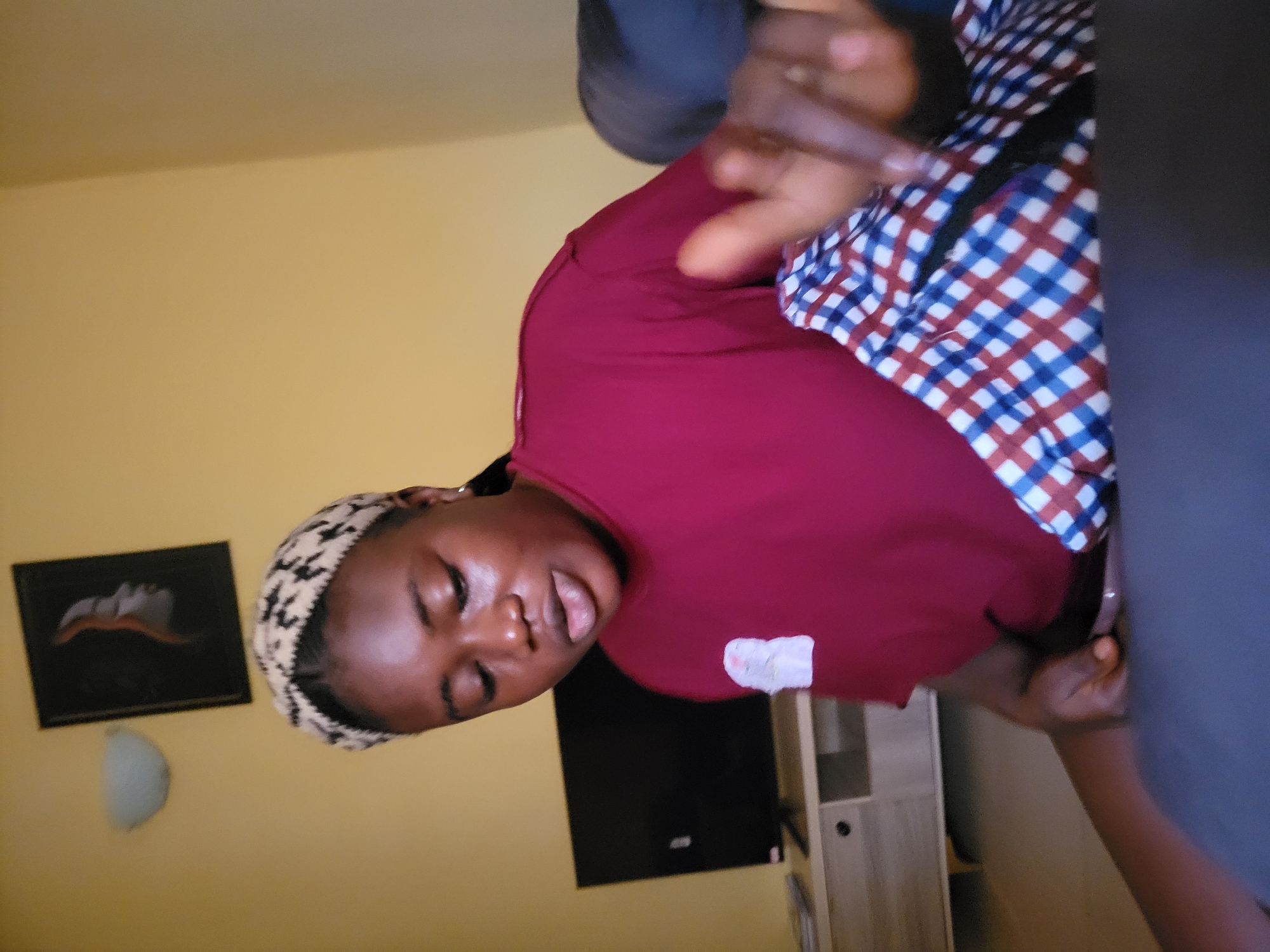 The image shows a young woman lying down in what appears to be a bedroom. She is wearing a maroon t-shirt and a headscarf with a black and white pattern. Her eyes are closed, and her mouth is slightly open, suggesting she might be sleeping or resting. Her right hand is raised, with fingers extended, and her left hand is resting on her chest.
Behind her, a wall is painted a light yellow color. On the wall to her left, there is a framed picture with a dark background and a light-colored abstract design. To her right, a television is visible, and next to it is a light-colored cabinet with shelves and drawers. The lighting in the room is dim, suggesting it might be evening or nighttime. The overall scene evokes a sense of privacy and relaxation. The location context provided is Suleja, Nigeria.