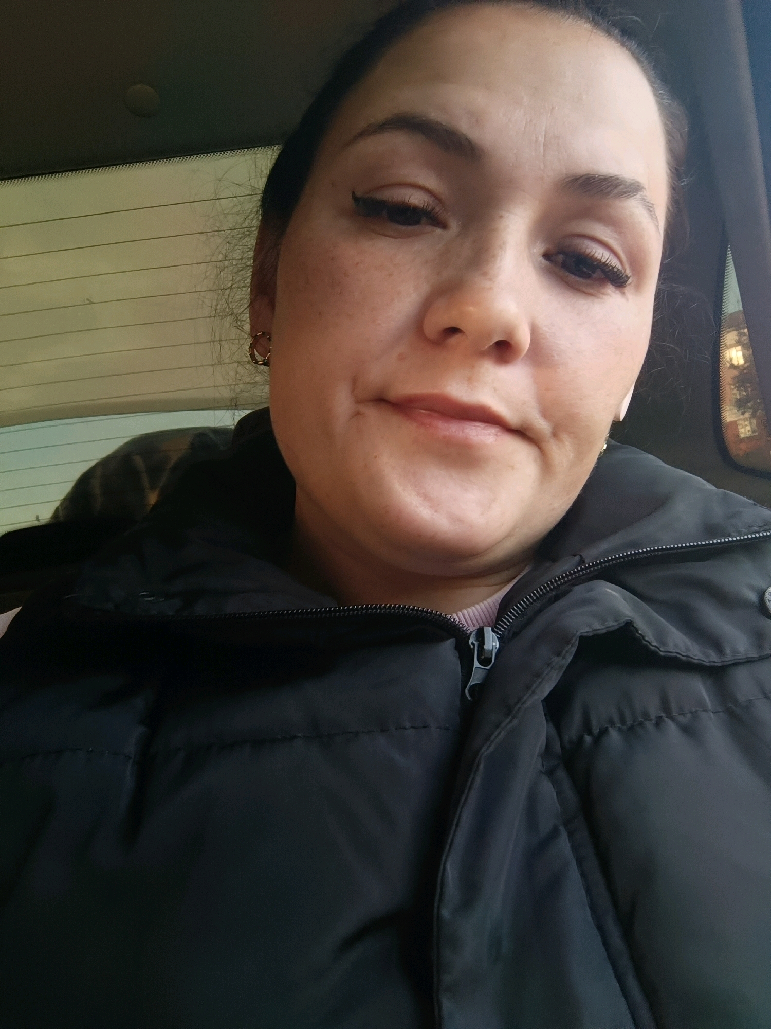 A close-up selfie shows a woman with brown hair, fair skin, and light brown eyes, wearing a black puffer jacket. She has freckles across her nose and cheeks, and she is wearing subtle eye makeup, including eyeliner and mascara, and a light lip color. She has on a small gold hoop earring in her left ear and appears to be wearing an earbud in her right ear. The background shows the interior of a vehicle, with a rear window and a dashboard visible. The lighting suggests it is daytime. It is not possible to determine the specific location within the city of Perm, Russia, based on the provided image.