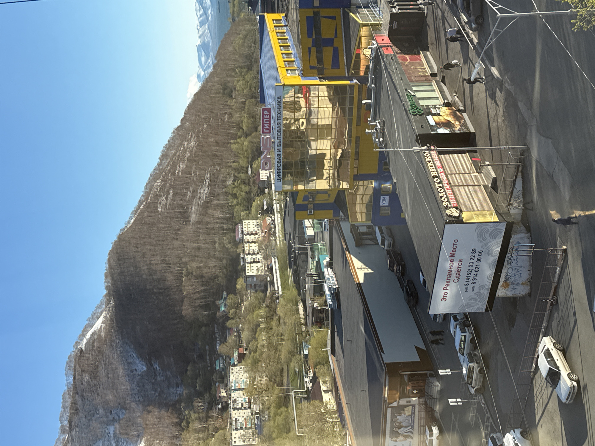 The image depicts a cityscape in Petropavlovsk-Kamchatsky, Russia. A mountain, partially covered in snow, rises in the background under a clear blue sky. In the foreground is a street with parked and moving vehicles. Notable buildings along the street include a blue and yellow building with a reflective glass facade, identified by a sign as "ГИПЕР Цифровая и Бытовая Техника" (Hyper Digital and Household Appliances). Another building is marked "Золото Золото ВиКKO" (Gold Gold VIKKO), likely a jewelry store. A billboard is visible, advertising a space for rent. Several people are seen walking along the street. The buildings suggest commercial activity in an urban environment.