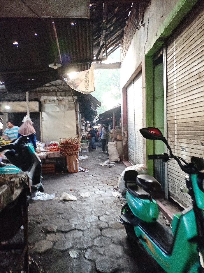 This image depicts a bustling, narrow alleyway within a traditional market in Mojosari, Indonesia. The scene is covered by makeshift roofing made of corrugated metal sheets and wooden beams, creating a partially shaded environment with pockets of natural light filtering through from open areas further down the path.
In the foreground on the right, a bright teal or turquoise scooter with a black seat and a distinctive red tail light integrated into its mirror is prominently parked. It stands next to a building featuring a light green-painted wall and a closed, white roll-down metal shutter. Another scooter is partially visible on the left foreground. The ground is paved with irregular, dark-colored stone or concrete blocks, which appear damp.
The midground reveals market activity. On the left, a stall displays produce, notably a large quantity of red chilies or peppers in woven baskets. Two individuals are present at this stall: one wearing a blue and white striped shirt, and another person whose head is covered with a light-colored veil or hijab. Further into the distance, the alley becomes more crowded with people and additional market stalls, although specific details are blurred. White sacks, possibly containing goods like rice, are visible on the ground near some stalls.
A single, bare light bulb hangs from the ceiling, providing localized illumination under the dark roof. A white fabric banner or sack, with partially discernible text such as "PAK" and "PADI" (likely referring to rice or a related product), is suspended from the roof structure. The time of day appears to be daytime, with a functional, rustic market aesthetic. No strong emotions are overtly displayed, but the scene suggests a regular day of commerce.