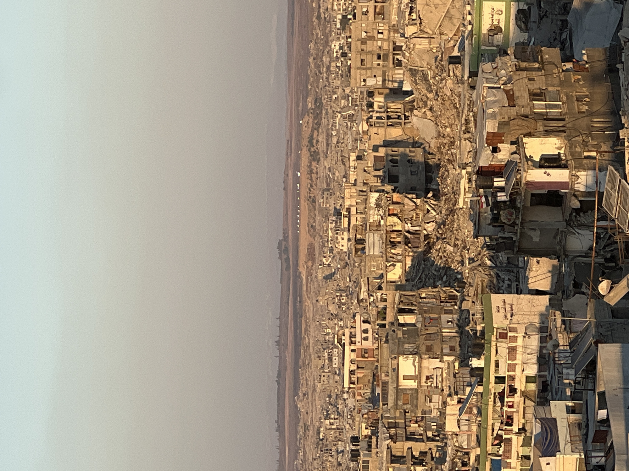 The image depicts a panoramic view of a densely populated urban area in the Gaza Strip, Palestinian Territories, characterized by widespread destruction and damage to buildings. The foreground and midground are filled with structures in various states of disrepair, ranging from partially collapsed walls and exposed rebar to completely ruined buildings reduced to rubble. Many buildings show signs of blast damage, with missing facades, shattered windows, and debris scattered around. Some structures appear to be partially standing, with internal walls and floors visible.
Interspersed among the damaged buildings are some that appear less affected, though they still show signs of wear and tear. These include multi-story apartment buildings with visible balconies, some adorned with laundry hanging to dry, and others with satellite dishes and water tanks on their rooftops. There are also makeshift structures, possibly temporary shelters, constructed from corrugated metal and tarpaulin.
The urban landscape stretches out towards the horizon, where more distant buildings and the surrounding terrain are visible. The ground appears to be a mix of sandy soil and the remnants of destroyed infrastructure. The sky above is clear and pale, with a gradient of light blue to a hazy beige near the horizon, suggesting either early morning or late afternoon light. The overall impression is one of significant devastation and the resilience of inhabitants in a conflict-affected zone. No people are clearly identifiable in the image.