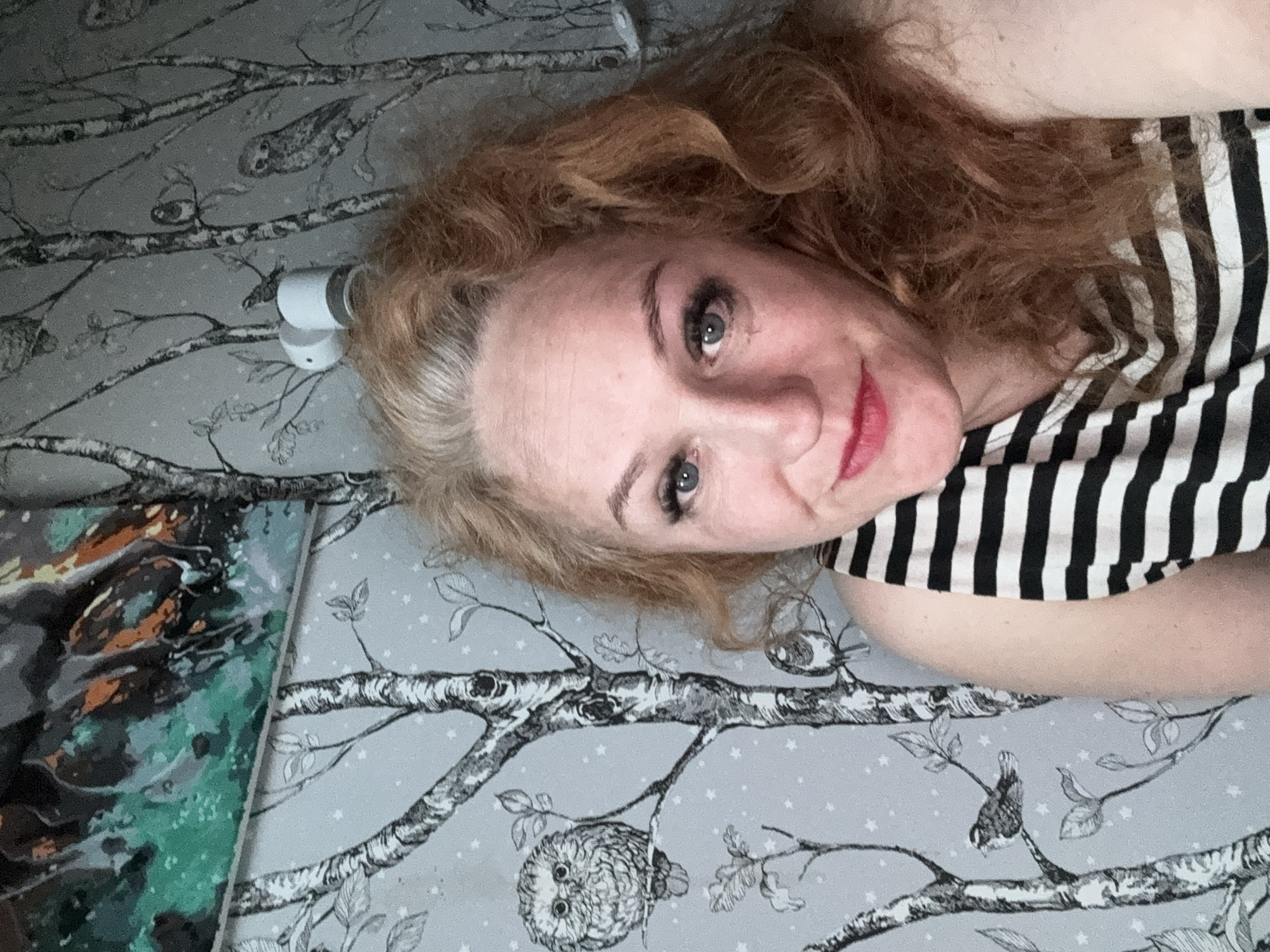 A woman with wavy, reddish-auburn hair and striking blue eyes is depicted lying down, looking directly at the camera with a gentle smile or smirk. Her hair shows some natural grey strands at the roots. She is wearing a black and white striped top.
The setting appears to be indoors, with the woman positioned on a light grey surface, likely bedding or a blanket, featuring a distinctive pattern. This pattern consists of stylized black and white tree branches, leaves, and various birds, including a prominent owl. Small white, star-like dots are scattered across the grey background.
To the left of the woman's head, partially obscured by her hair, a small white cylindrical object is visible, possibly a personal device or part of the room's fixtures. In the lower-left corner of the frame, a portion of an abstract or artistic print, characterized by shades of green, brown, and black, is visible. The image conveys a relaxed and personal atmosphere. No text is visible. The context provided for the image is Chaikovsky, Russia.