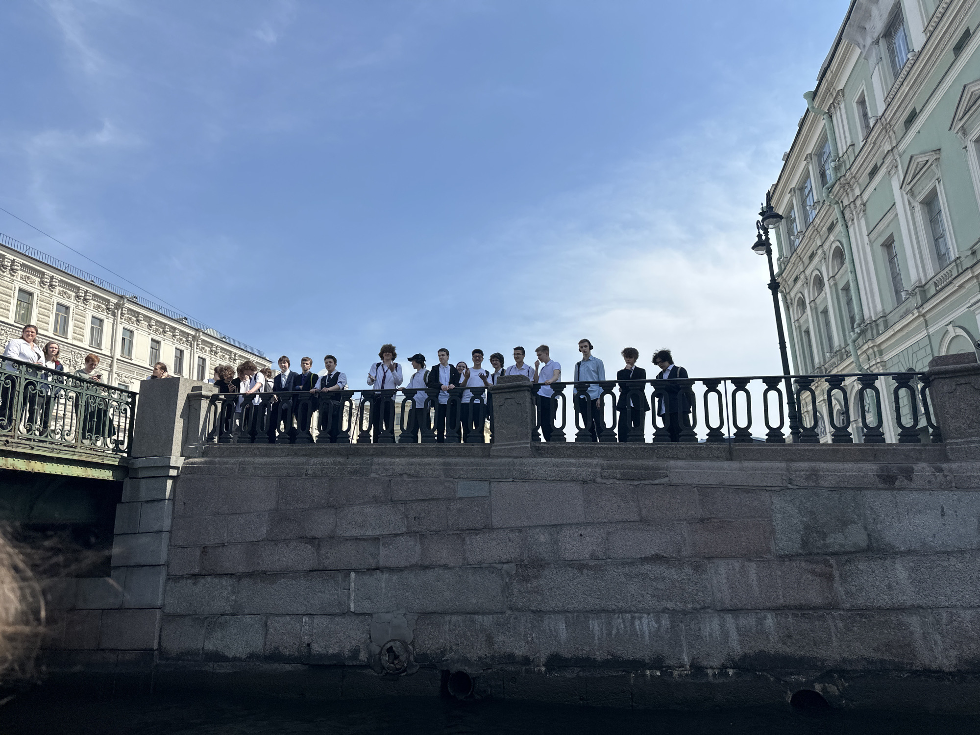 A wide-angle shot taken at water level captures a stone embankment in Saint Petersburg, Russia. A group of about twenty young people, mostly males, are standing along the decorative black cast-iron fence that tops the wall. They appear to be students; several are wearing white shirts, and some have ties or dark jackets. They are likely graduating students.
To the left, other individuals are standing on a bridge, and on the right, a pale-green and white building with ornate architectural details rises vertically. A black lamppost stands in front of the building. The sky is mostly clear with some wispy clouds. The water surface is dark. A hint of hair is visible in the foreground.