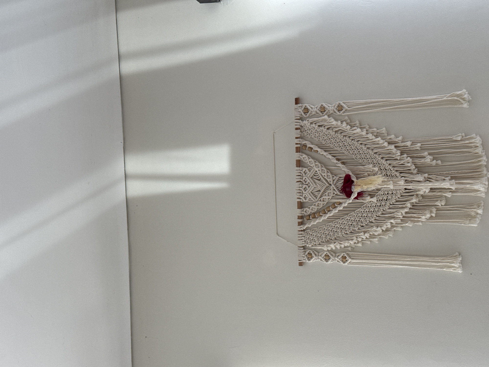 A macramé wall hanging with white cords and wooden beads, adorned with a red and tan tassel in the center, is mounted on a light-colored wall. The wall is illuminated by diffused light with visible shadows cast from an unseen source above. The location is possibly Kuta Selatan, Indonesia, based on implied information. There are no people present in the image.