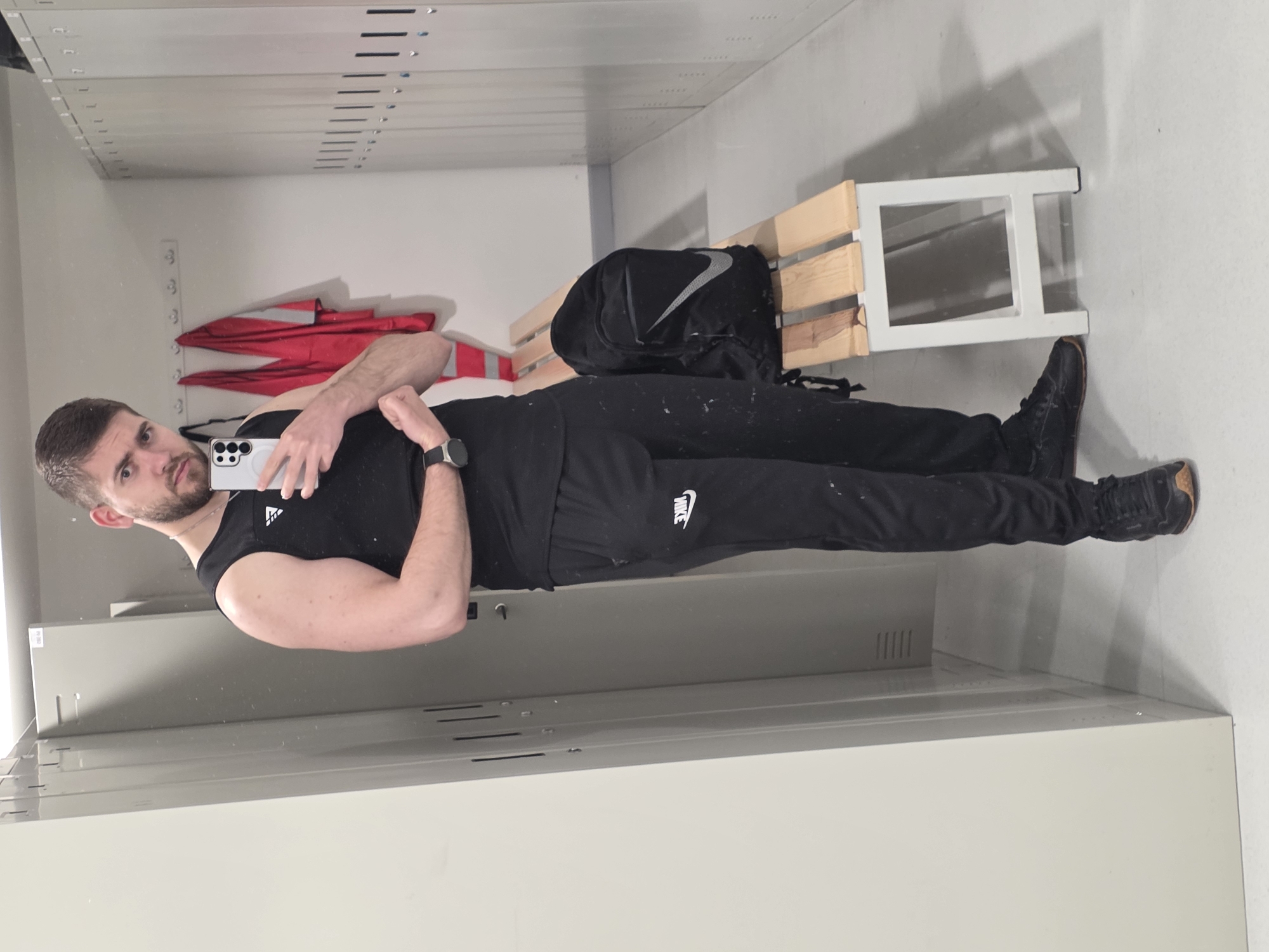 The image, set in a changing room in Plovdiv, Bulgaria, captures a man taking a mirror selfie. He stands upright, facing the camera with a neutral expression. The man has short, dark hair and a beard, and is wearing a black sleeveless top with an "adidas" logo on the left chest, black track pants featuring a white "NIKE" logo on the left thigh, and dark athletic shoes. A dark smartwatch is visible on his left wrist, and he holds a light-colored Samsung smartphone in his left hand to take the photo.
The background shows a functional indoor space, likely a gym or workplace locker room. The walls are light-colored, and rows of grey metal lockers with horizontal ventilation slits are prominent on both sides of the frame. Behind the man, a wooden slatted bench with a white frame holds a black backpack adorned with a large white Nike swoosh logo. A folded red jacket hangs on a hook behind the bench. The lighting is even, typical of an indoor environment, with no discernible time of day or specific weather indicated. The overall scene suggests a moment before or after physical activity.