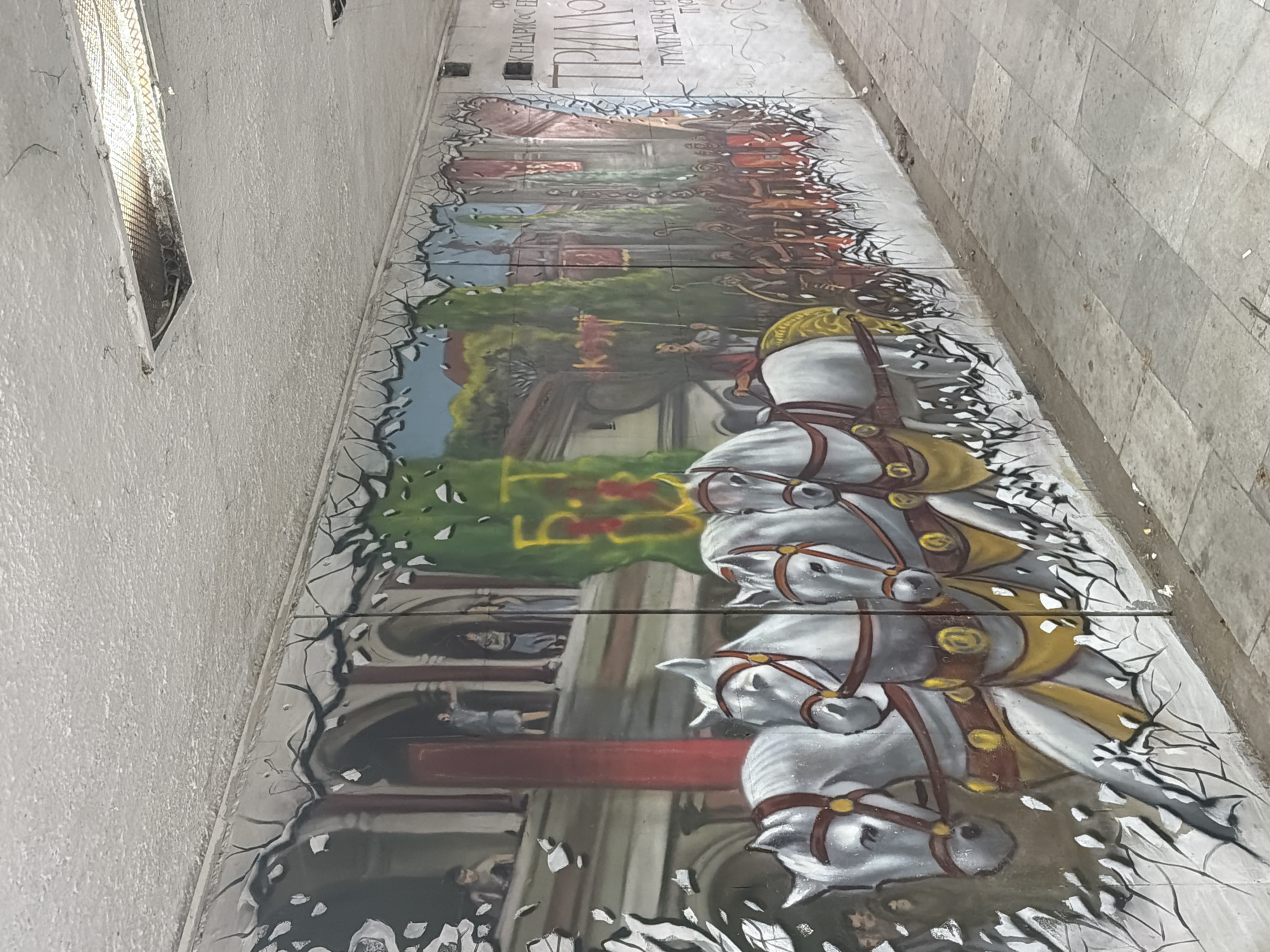 The image shows a mural depicting a historical scene, likely related to the city's history. The mural is painted on a long, narrow surface, possibly a sidewalk or the side of a building. The scene depicts a procession of people on horseback, with architectural elements such as arches and columns in the background. The mural is stylized with some portions looking like they are cracked and falling apart. The location is likely in the city of Plovdiv, Bulgaria. The mural is on the ground, where it is a section of sidewalk.