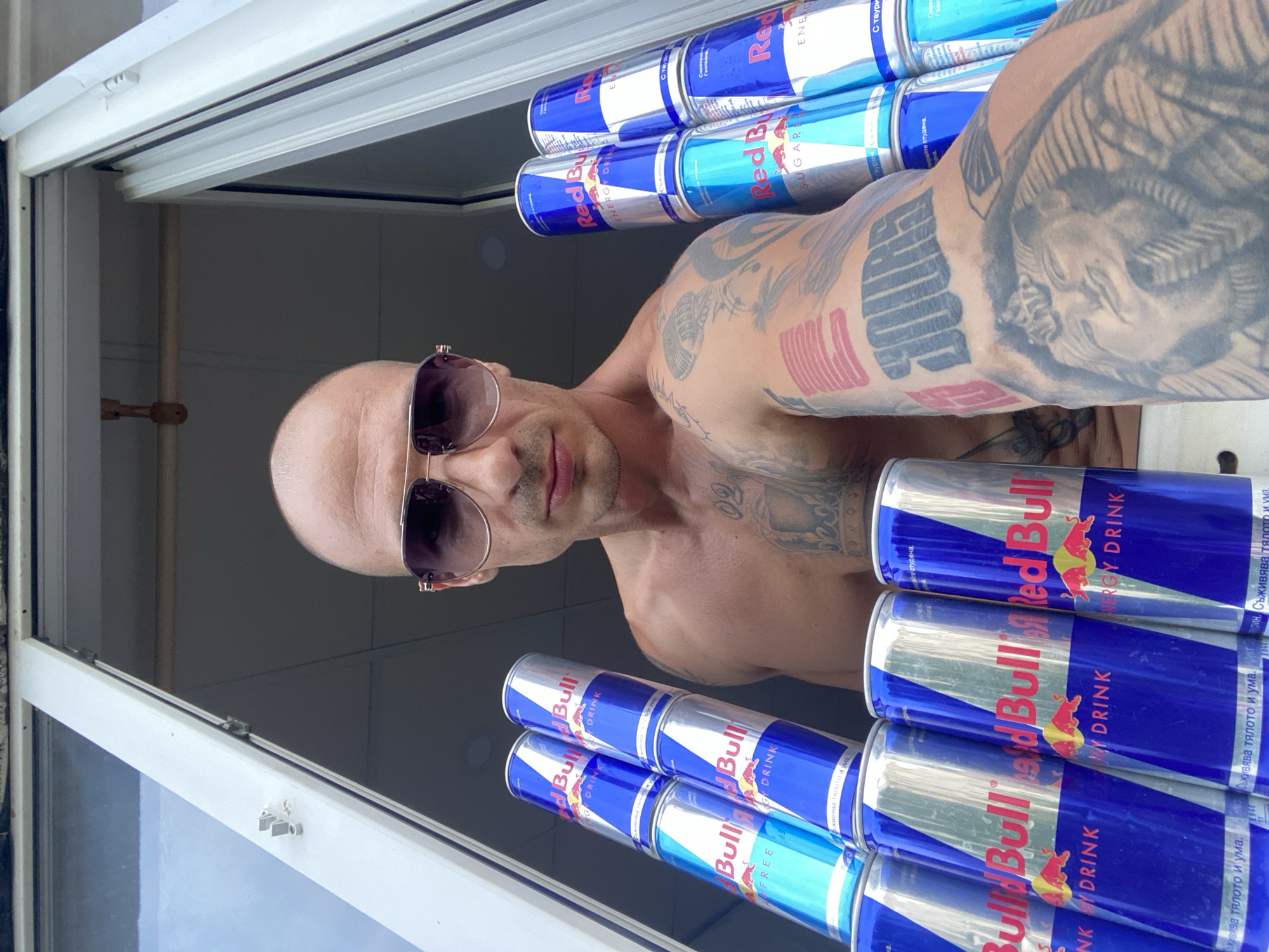 A shirtless man with a shaved head and sunglasses is holding several cans of Red Bull. He has tattoos on his arm and chest, including a pharaoh's head on his bicep. The man is positioned in front of an open window, suggesting he is either indoors looking out or outdoors looking in. The background appears to be a grey, indistinct interior space. The Red Bull cans are stacked in multiple piles. This image was taken in the city of Pleven, Bulgaria.