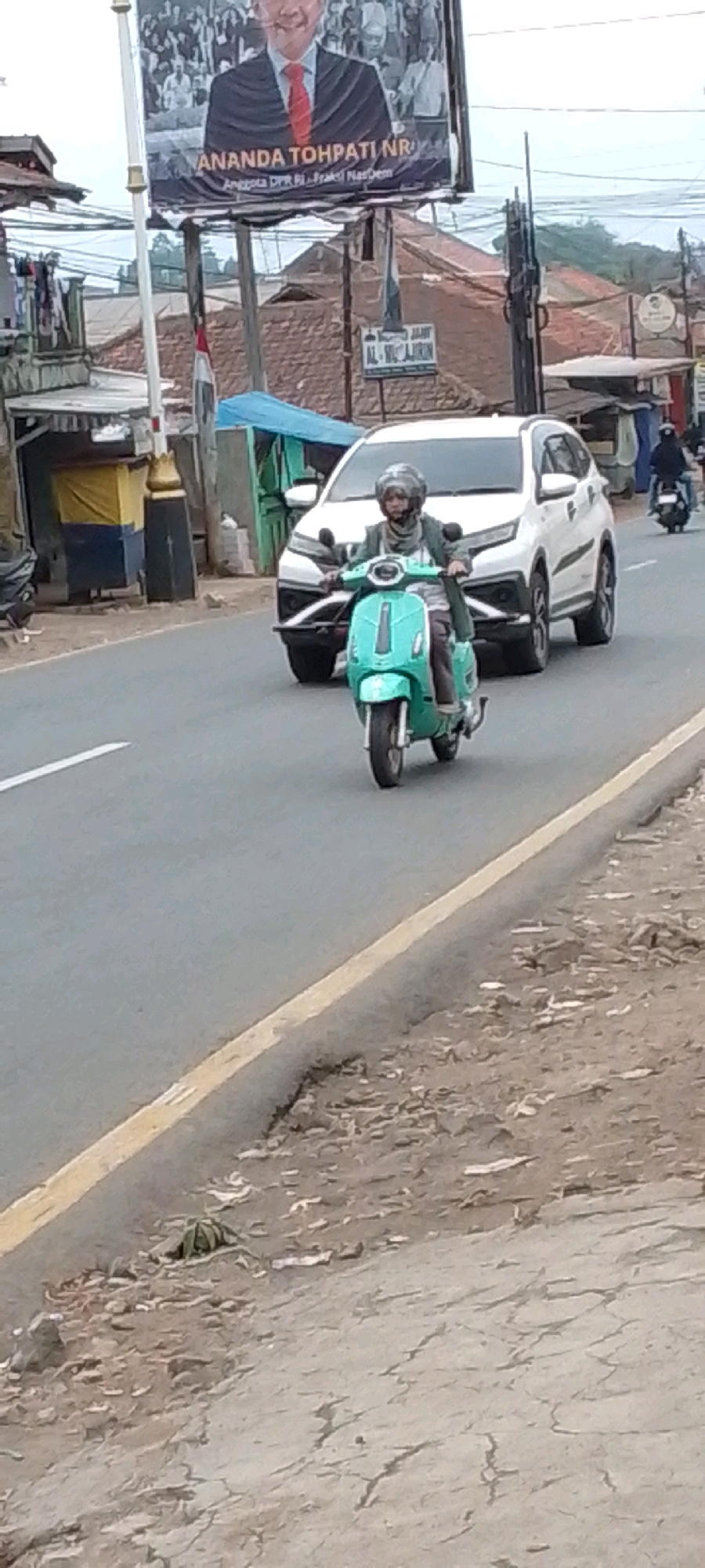 The image captures a street scene in Cilaku, Indonesia, with a focus on a person riding a turquoise scooter. The scooter rider, wearing a grey headscarf and a dark jacket, is positioned centrally in the frame, facing the viewer. Behind the scooter, a white SUV is visible, indicating traffic on the road. In the background, a large billboard featuring a man in a suit and tie with text in Indonesian is prominently displayed. The environment appears to be an urban or semi-urban setting with buildings and vegetation visible. A yellow line marks the edge of the road.