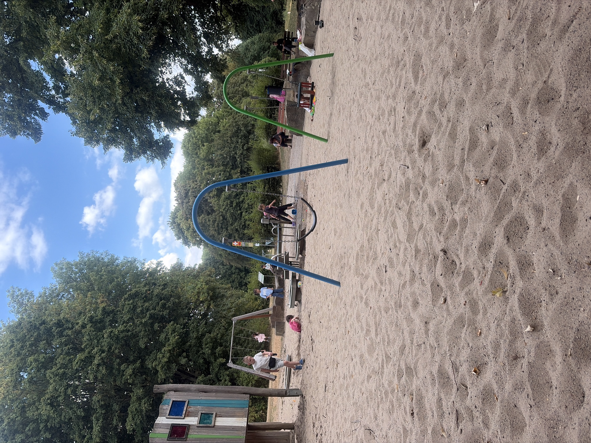 The image depicts a children's playground in Duisburg, Germany, featuring a sandpit with several swings. In the foreground, a young boy in a white striped shirt and black shorts walks across the sand. To his left, a child in a pink shirt is lying on the sand. Further back, two girls are on a swing set, one in a pink dress and the other in blue. Another child in a blue shirt stands near a wooden structure with colorful panels. Two adults are seated on a bench in the background, and other adults are visible further back, near trees and more play equipment. The playground is surrounded by lush green trees under a partly cloudy blue sky.