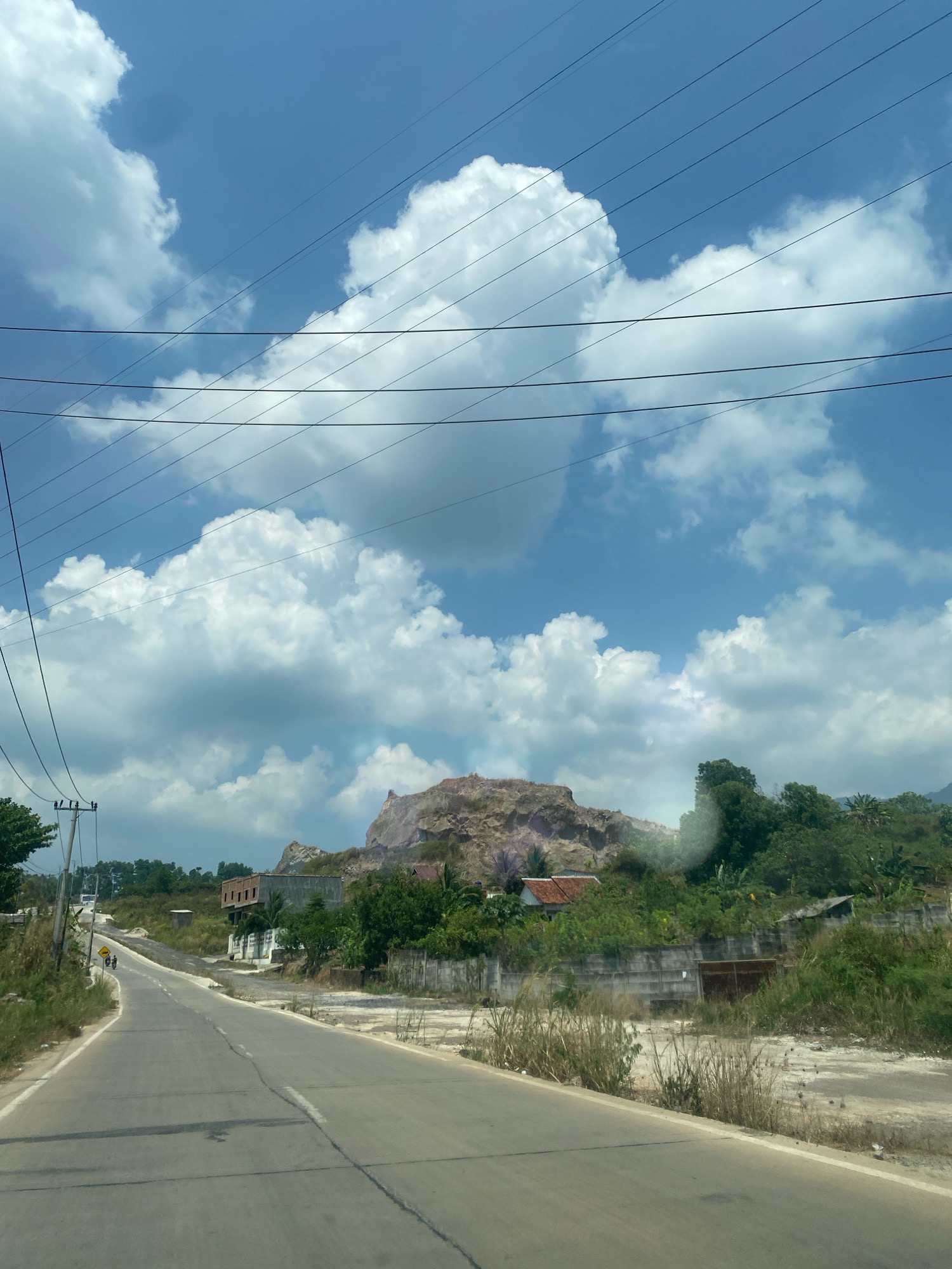 The image depicts a rural roadside scene in Bandar Lampung, Indonesia, under a partly cloudy blue sky. A paved road curves to the left, with dry grass and some scattered debris along its right side. On the left side of the road, there is a utility pole and some vegetation. In the background, there is a large, rocky hill with evidence of quarrying or erosion, showing exposed, reddish-brown rock formations. Several buildings, including a multi-story concrete structure with visible windows and a house with a terracotta-tiled roof, are nestled amongst lush green trees and vegetation on the slopes leading up to the hill. Power lines stretch across the upper portion of the image. Two individuals on a motorcycle are visible on the road in the distance. The overall impression is of a somewhat undeveloped area on the outskirts of a city, with a mix of natural landscape and human habitation.
