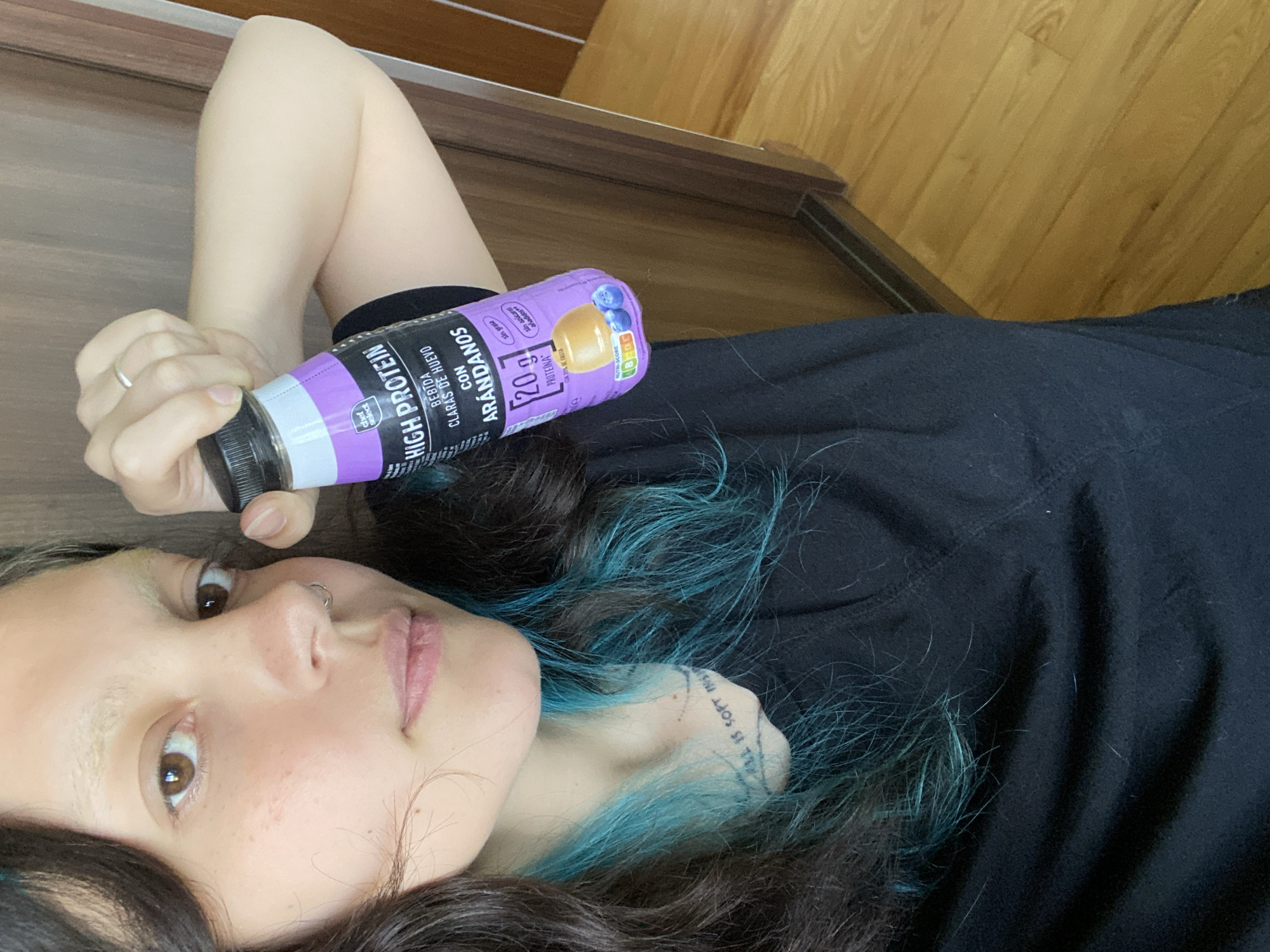 Here's a detailed description of the image:
**Scene:** A selfie taken from a lying down position. The subject is a person with dark hair that is highlighted with teal/blue streaks. They are wearing a black top. They appear to be indoors, on a brown hardwood floor, adjacent to a vertical wooden-paneled wall.
**People:** The primary subject is a person lying down. Their face is partially visible, showing light skin, brown eyes, and a nose ring. A tattoo is visible on their wrist.
**Objects:** The subject is holding a purple bottle labeled "High Protein," with Spanish text including "Arandanos" (blueberries). The bottle contains 20g of protein, and a description of it being made with egg whites and blueberries.
**Actions/Interactions:** The person is holding the protein drink. They are looking at the camera.
**Environment:** The photo appears to be taken inside a room with hardwood floors and wooden walls.
**Location:** It is NOT possible to determine if this image was taken in Villaviciosa, Spain based solely on visual cues. There are no distinguishable landmarks or details in the photo that would pinpoint the location.