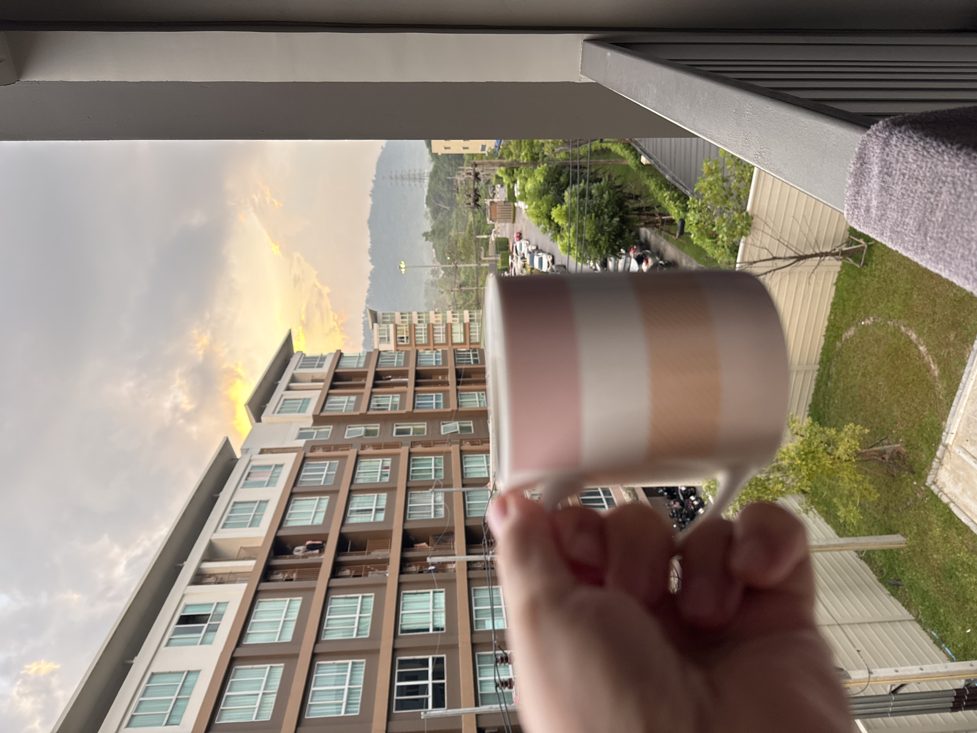 In Kathu, Thailand, at dawn, a hand holds a mug with horizontal pink and beige stripes, the rim of which is in the foreground. The mug is held up to a balcony, overlooking an apartment building with brown and white facades and numerous windows. Beyond the building, a street is visible, lined with parked cars and lush green trees. In the distance, rolling hills are shrouded in a misty haze, and a power pylon stands prominently against the cloudy sky, which is illuminated by the soft glow of sunrise. A lavender towel is draped over the balcony railing in the lower right corner of the frame.