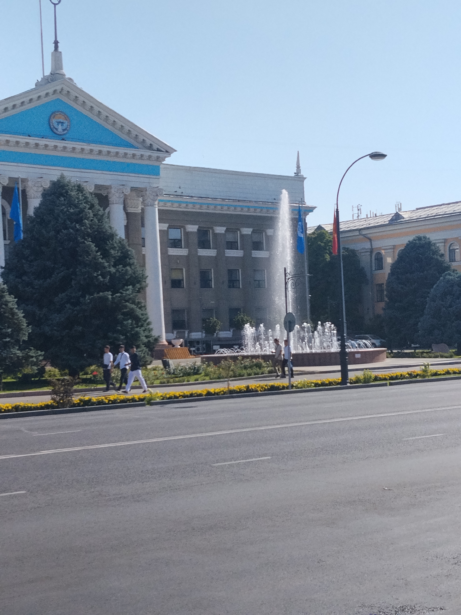 The image shows the Ala-Too Square in Bishkek, Kyrgyzstan, on a bright, sunny day. In the foreground is a wide, empty asphalt road with white lane markings. Beyond the road is a sidewalk lined with yellow flowers and green bushes. Behind the flowers is a large fountain with several tiers of spraying water. To the left of the fountain is a large, imposing building with white columns and a blue triangular pediment adorned with a circular emblem. Two blue flags are visible on the building. Several people are walking on the sidewalk in front of the fountain and building. One of the people appears to be wearing a white shirt and dark pants, while another is wearing a white shirt and light-colored pants. To the right of the fountain and building, another building with yellow accents is visible, partially obscured by trees. A lamppost with a curved arm stands near the fountain. The sky is a clear, pale blue.