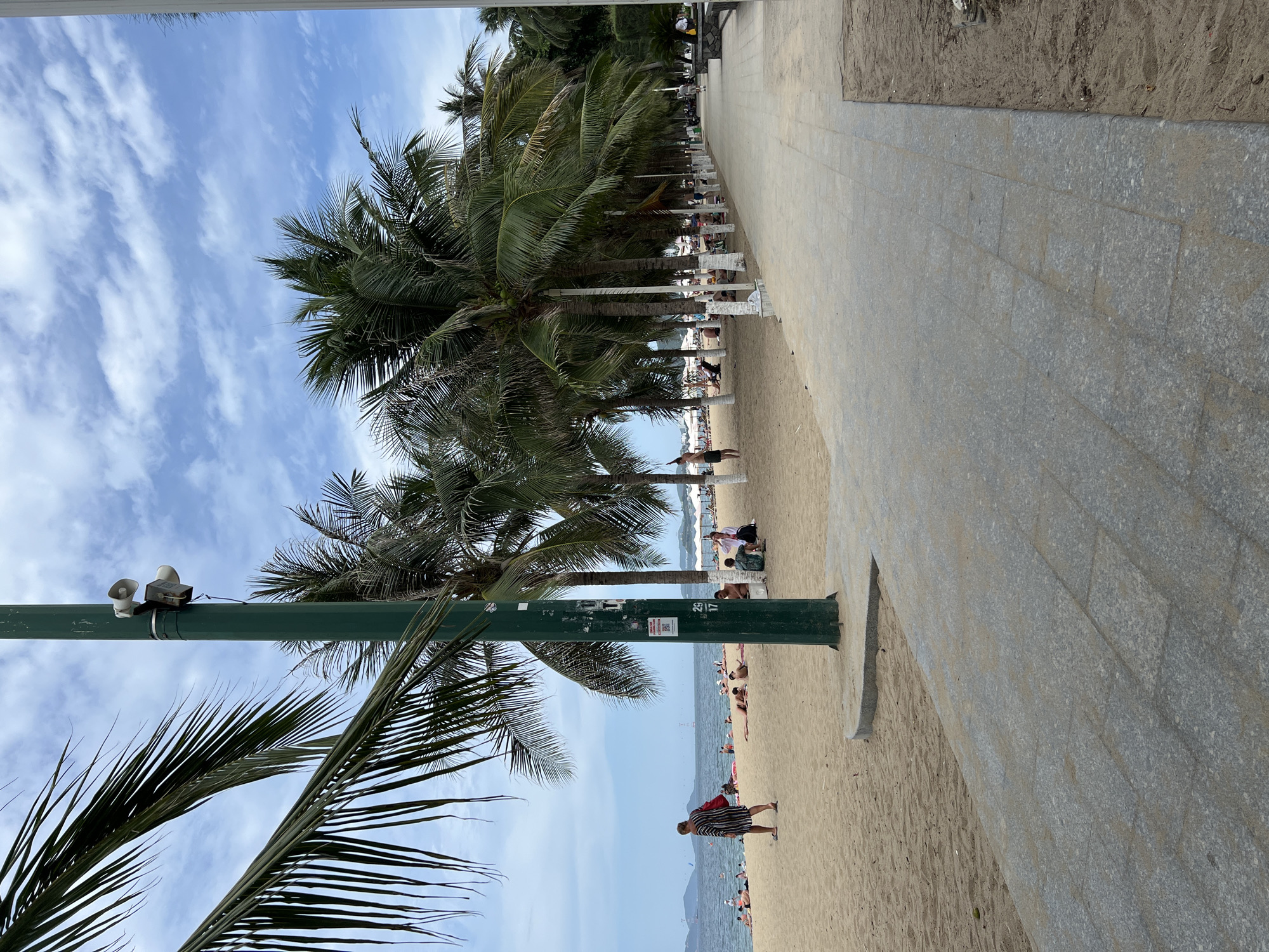 The image depicts a paved walkway alongside a sandy beach in Nha Trang, Vietnam. Palm trees line the edge of the walkway, casting shadows on the sand where numerous beachgoers are scattered. Some individuals are sunbathing, while others are seated or walking. In the distance, the ocean is visible with a cloudy sky overhead. A prominent green pole with speakers and a poster is located on the left side of the walkway, near the foreground. The walkway is constructed of large, grey stone pavers. The scene conveys a relaxed, tropical atmosphere.