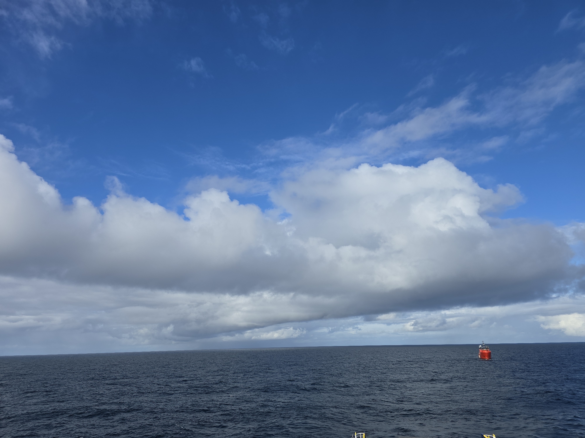 The image depicts a seascape under a partly cloudy sky. The sea is dark blue and stretches to the horizon. A red buoy or small boat is visible in the distance on the right side of the frame. The sky is mostly blue with patches of white and grey clouds. The location is Unknown, Unknown.