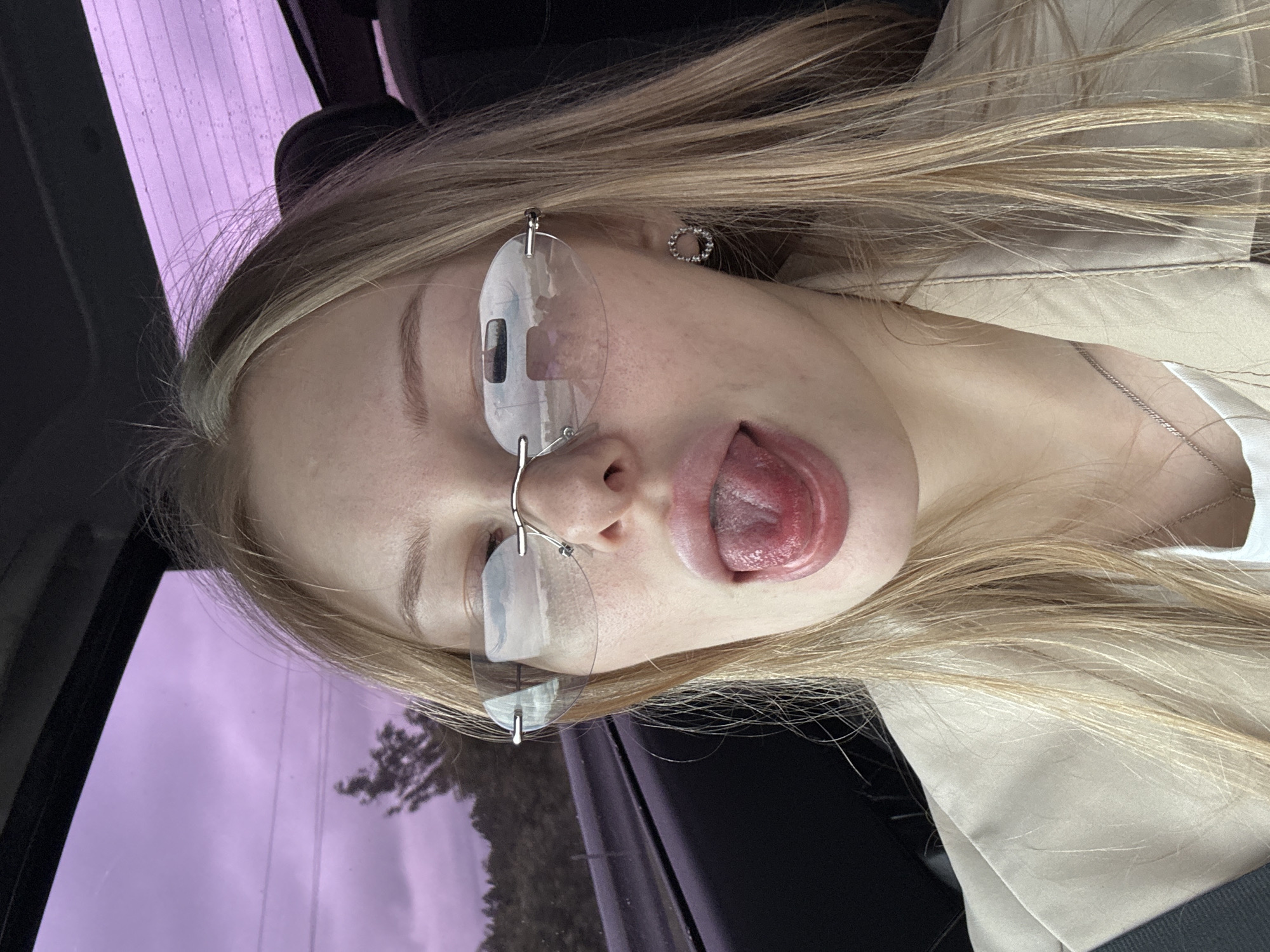 A young woman with blonde hair is pictured inside a vehicle, with her tongue sticking out. She is wearing rimless sunglasses and a light-colored jacket over a white top. A silver necklace is visible around her neck. The background through the car windows shows a blurry landscape of trees and a cloudy, purple sky, suggesting it is daytime. The interior of the car is dark and out of focus, with only the woman and her immediate surroundings clearly visible. The location is identified as Kondratovo, Russia.
