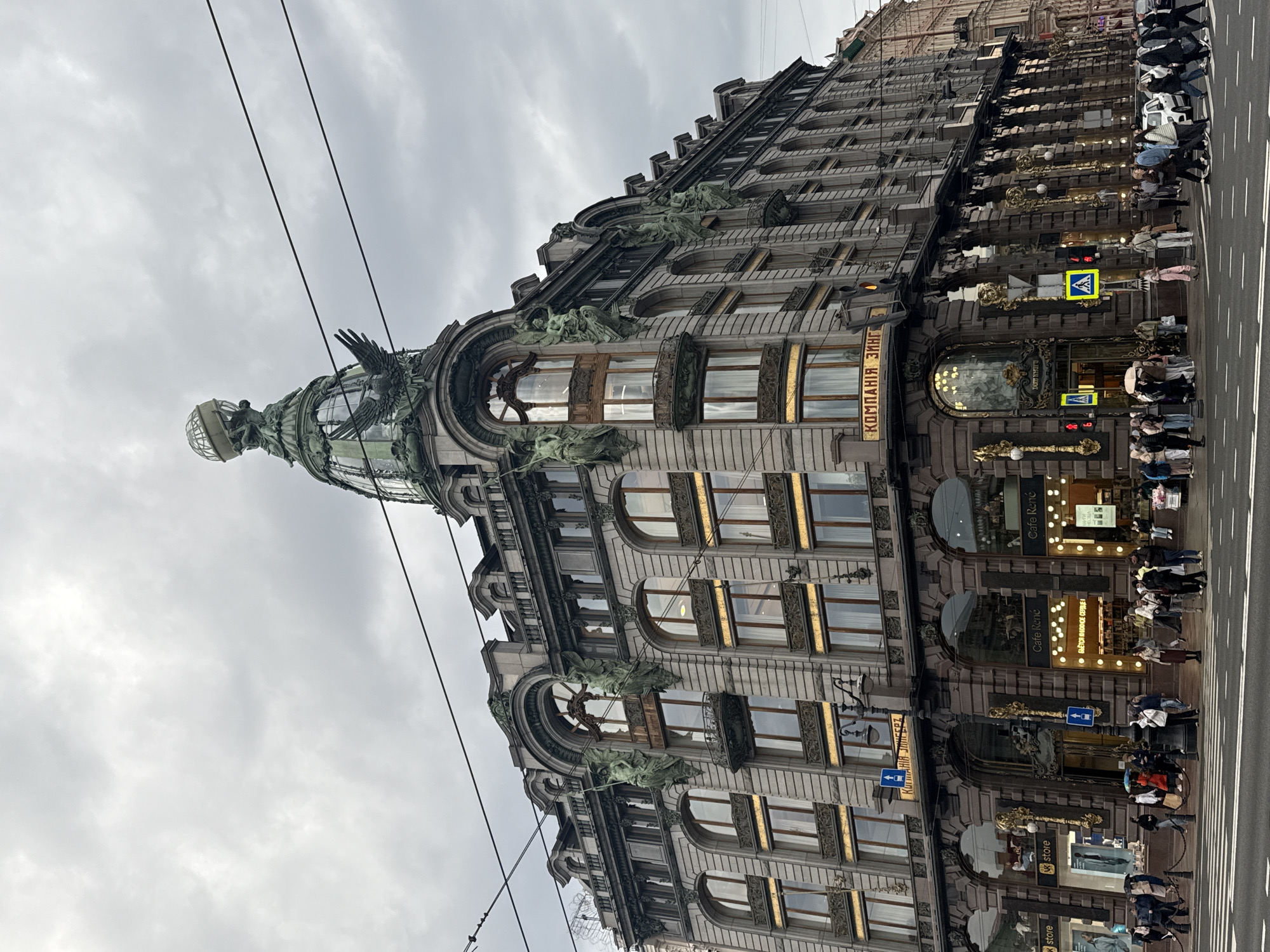 The image captures a street-level view of the historic Singer House, now known as Dom Knigi (House of Books), in Saint Petersburg, Russia. The building's ornate facade, with its distinctive glass dome, sculptures, and architectural details, is prominently displayed. The name "КОМПАНІЯ ЗИНГ" (Company Zing) is visible on the building. People are seen walking and crossing the street in front of the building. There are commercial businesses and shops visible in the building's lower levels. The sky is overcast, suggesting a cloudy day.