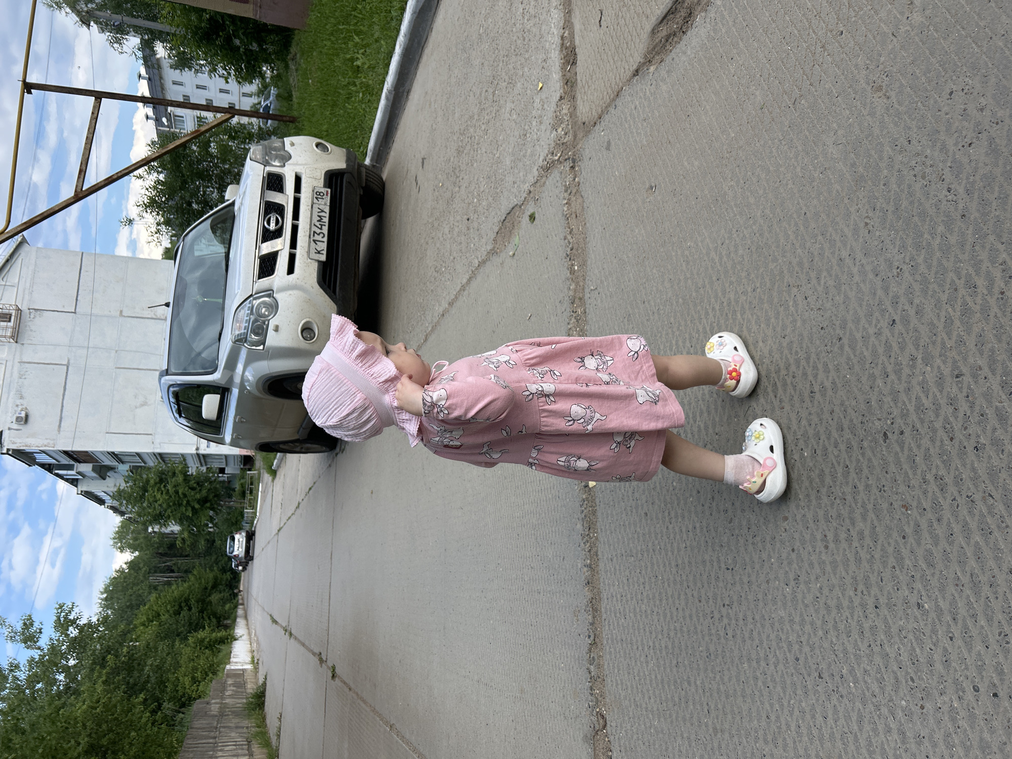 A young girl wearing a pink dress and a pink bonnet stands on a concrete path. She has a toy crown on her left shoe and a flower charm on her right shoe. Her pink dress features a pattern of white rabbits. The girl is looking away from the camera, towards a white Nissan SUV parked on the right side of the street. The SUV has a license plate that reads "K134MY 18." In the background, there is a multi-story building and a line of trees. The scene appears to be an urban street in Noviy, Russia.