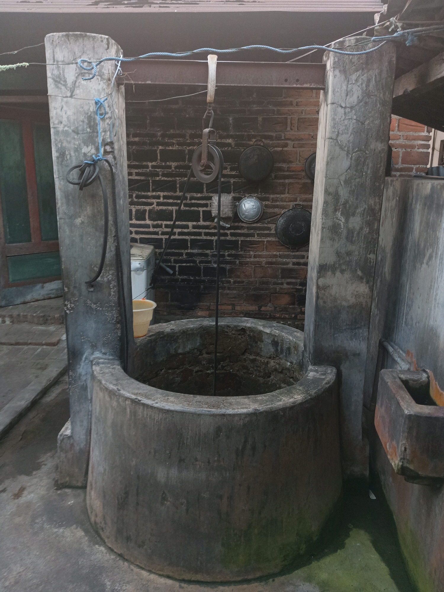 An outdoor traditional well is depicted in the village of Ngrendeng, Indonesia. The well structure consists of a circular concrete basin, surrounded by two large, weathered concrete pillars. A pulley system, suspended from a horizontal bar, is positioned above the well opening. A rope extends from the pulley into the well. The background features a brick wall with several metal discs or pots hanging on it. To the left of the well, a light-colored plastic bucket is visible. The ground around the well is paved and appears damp in places, with some green algae growth. To the right of the well, there is a rectangular concrete structure, possibly a basin or trough. The overall scene suggests a utilitarian setting, likely for water collection. The lighting is dim, indicating it might be dusk or an overcast day. No people are present in the image.