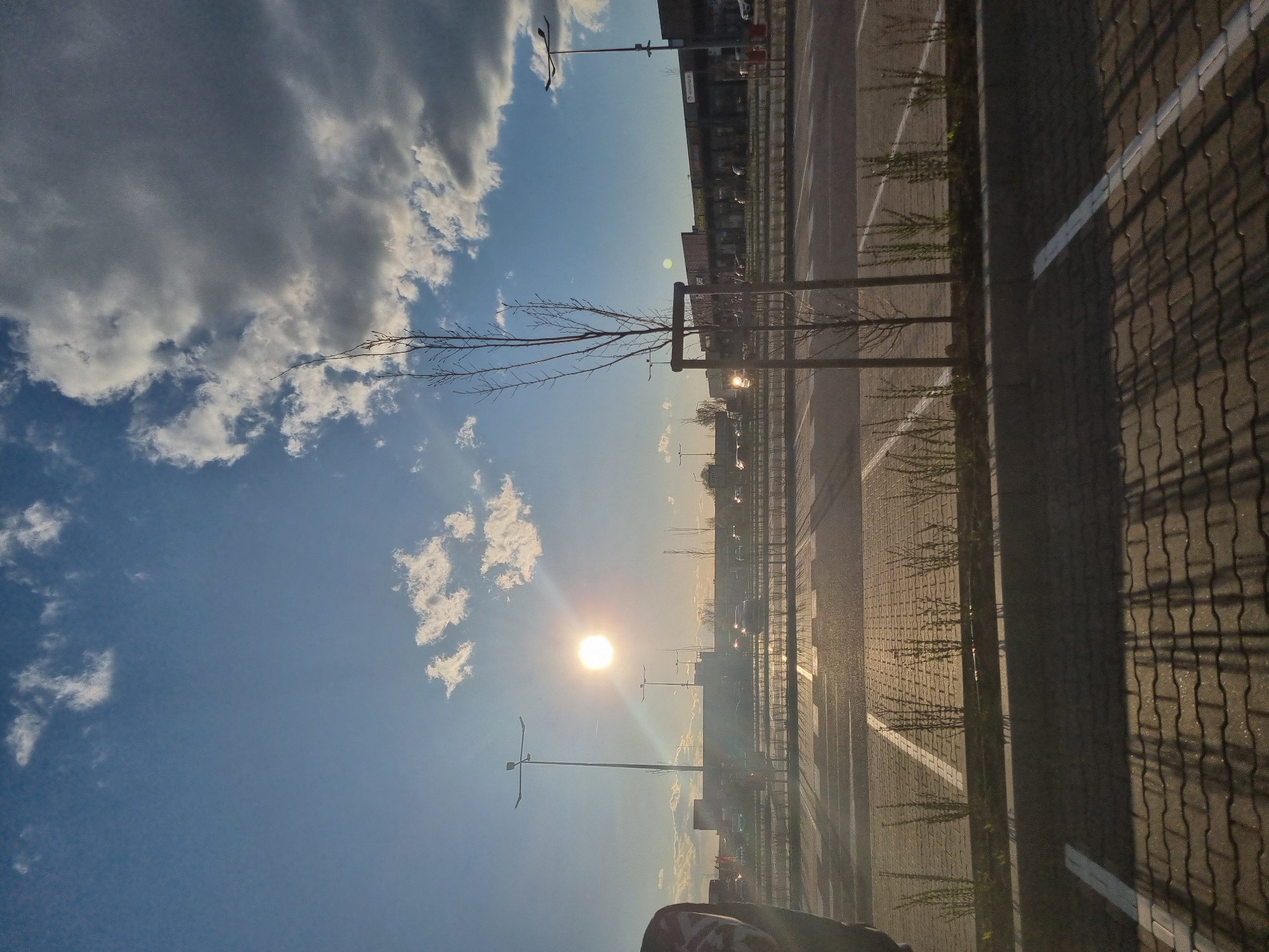 The image depicts a sunlit urban landscape, likely a parking lot in Sofia, Bulgaria. The sky is partly cloudy with the sun shining brightly. In the foreground are parking spaces demarcated by white lines, with trees planted along the curb. A metal fence casts a shadow over the paved surface. In the distance, buildings and streetlights are visible. There is a hint of a person visible in the bottom part of the image.