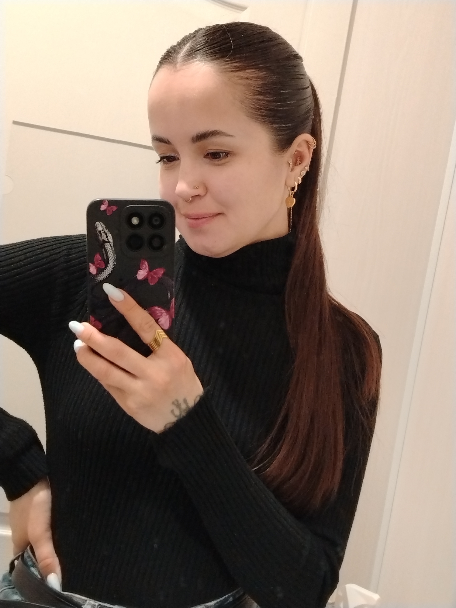 A woman is taking a selfie in what appears to be an indoor location, possibly a bathroom, given the white door in the background. She has brown hair pulled back into a ponytail and is wearing a black turtleneck sweater. She is holding a black smartphone with a decorative case featuring a snake and butterflies. She has multiple piercings in her left ear and a nose ring. A gold ring is visible on her finger. Her fingernails are painted white. Part of her jeans is visible where her left hand is positioned. There is no information to confirm the image was taken in Chaikovsky, Russia.
