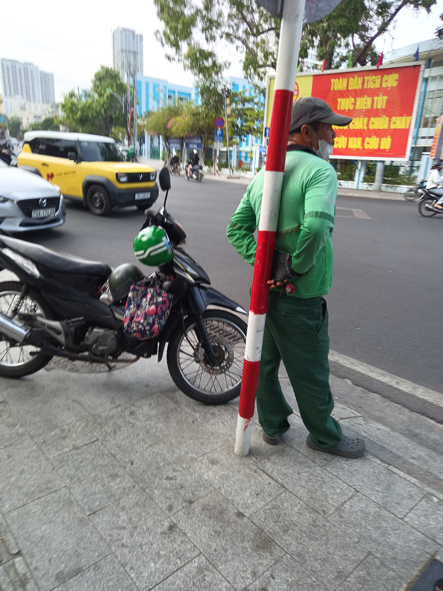 The image shows a man wearing a green long-sleeved shirt, green pants, a gray baseball cap, and a white face mask, holding a red and white striped pole. He is standing on a sidewalk next to a black motorcycle with a green helmet and a floral bag attached to it. In the background, there is a yellow SUV, other motorcycles, a large red banner with Vietnamese text, buildings, and trees. The scene appears to be a street in Nha Trang, Vietnam, given the presence of Vietnamese text and the common modes of transportation. The man's posture and the pole suggest he might be a street vendor or a worker directing traffic or performing some roadside service.