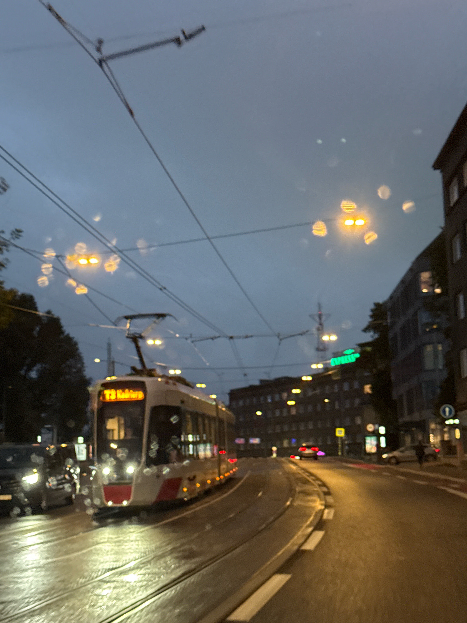 This is a slightly blurred, low-light photograph taken from the perspective of someone inside a vehicle, likely a tram or car, looking forward along a wet city street. The scene appears to be Tallinn, Estonia, given the tram's display and the architecture.
Dominating the foreground are tram tracks on a curving road. A modern tram, white with red accents, is on the tracks, its front illuminated. The tram's digital display shows "T3 Ratasepa." The street and tram tracks are wet, reflecting the lights. To the left, a dark car is visible, its headlights on.
In the background, buildings line the right side of the street, appearing to be a mix of older and newer constructions with visible windows and some illuminated interiors. Streetlights and building lights cast a yellow and white glow, interspersed with circular bokeh effects, suggesting the photograph was taken through a wet window. Overhead, a network of tram wires crisscrosses the sky, silhouetted against a dark, overcast sky. Some of the lights in the distance are blurred green neon signs. The overall atmosphere is one of evening or dusk in an urban environment, possibly during or after rain.