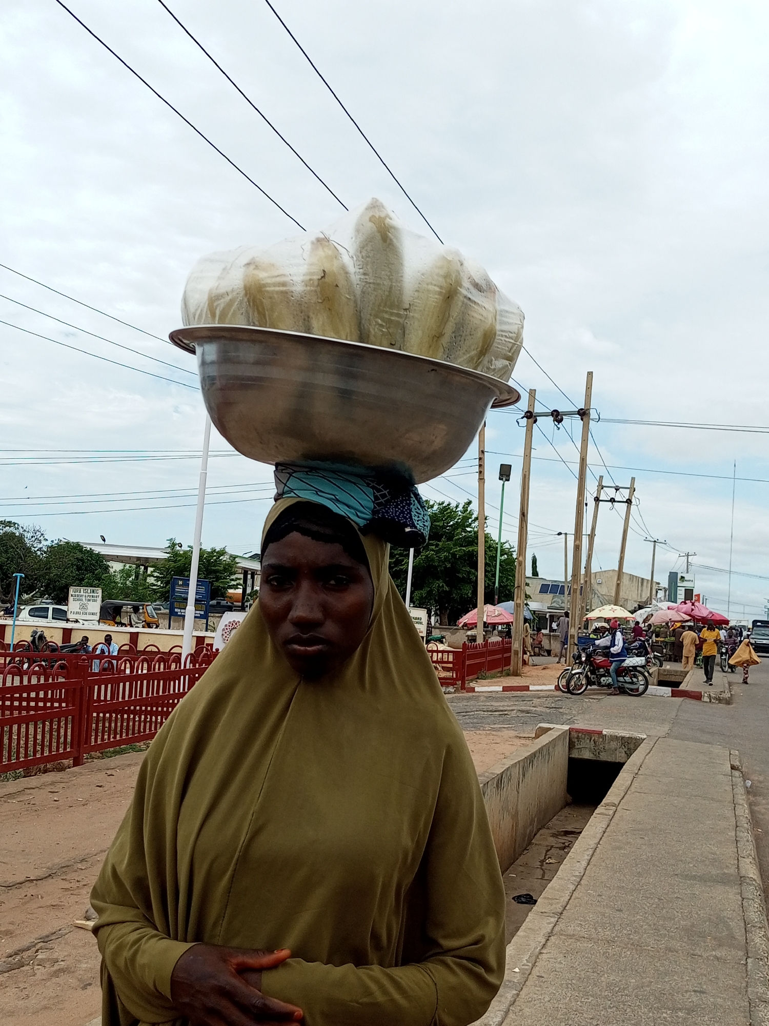 The image shows a woman carrying a large metal bowl filled with wrapped items, possibly food, on her head. She is wearing a green hijab and is standing on a street in Tempure, Nigeria. The background shows a street scene with several stalls, people, and vehicles, including motorcycles. There are also power lines and utility poles visible. The woman appears to be a street vendor.