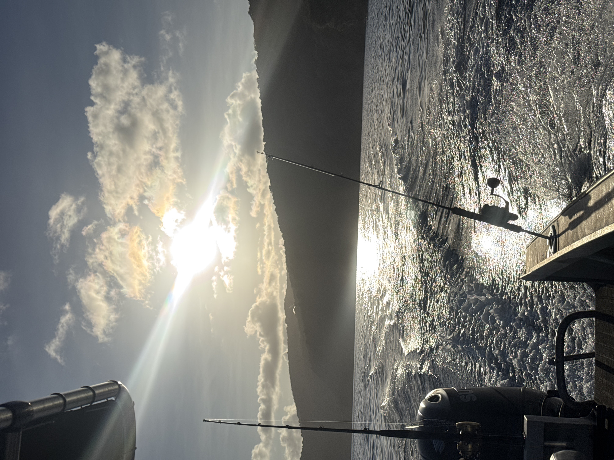 The image shows a boat moving through the water, creating a wake. There are two fishing rods visible, one on each side of the boat. The sun is setting behind a silhouetted island, casting a warm glow over the scene. The water is choppy, and the sun's rays are reflecting off the surface, creating a shimmering effect. The island is covered in lush vegetation, and there are clouds in the sky. The boat appears to be a fishing boat, with an outboard motor visible at the stern. The overall impression is one of a peaceful and serene day at sea.