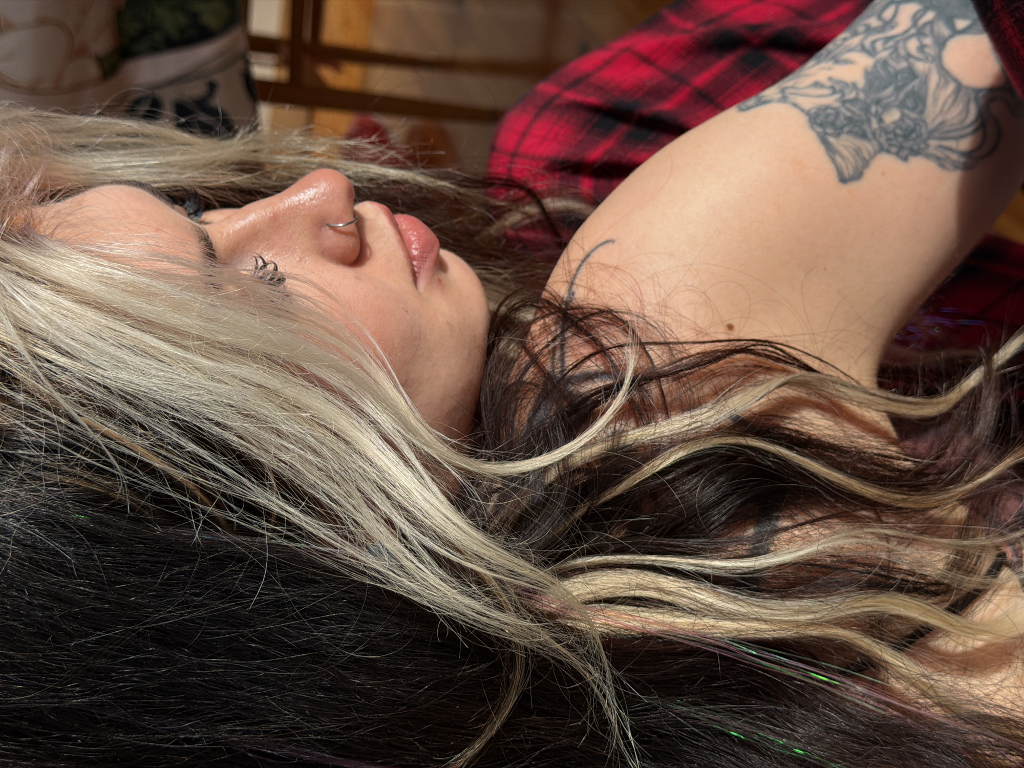 A woman with a fair complexion, blonde-streaked hair, and multiple piercings in her nose and ear lies supine on a surface. She has a tattoo on her left arm. The backdrop includes a plaid fabric and a wooden structure. The location and city of Budva, Montenegro are not determinable from the provided image.