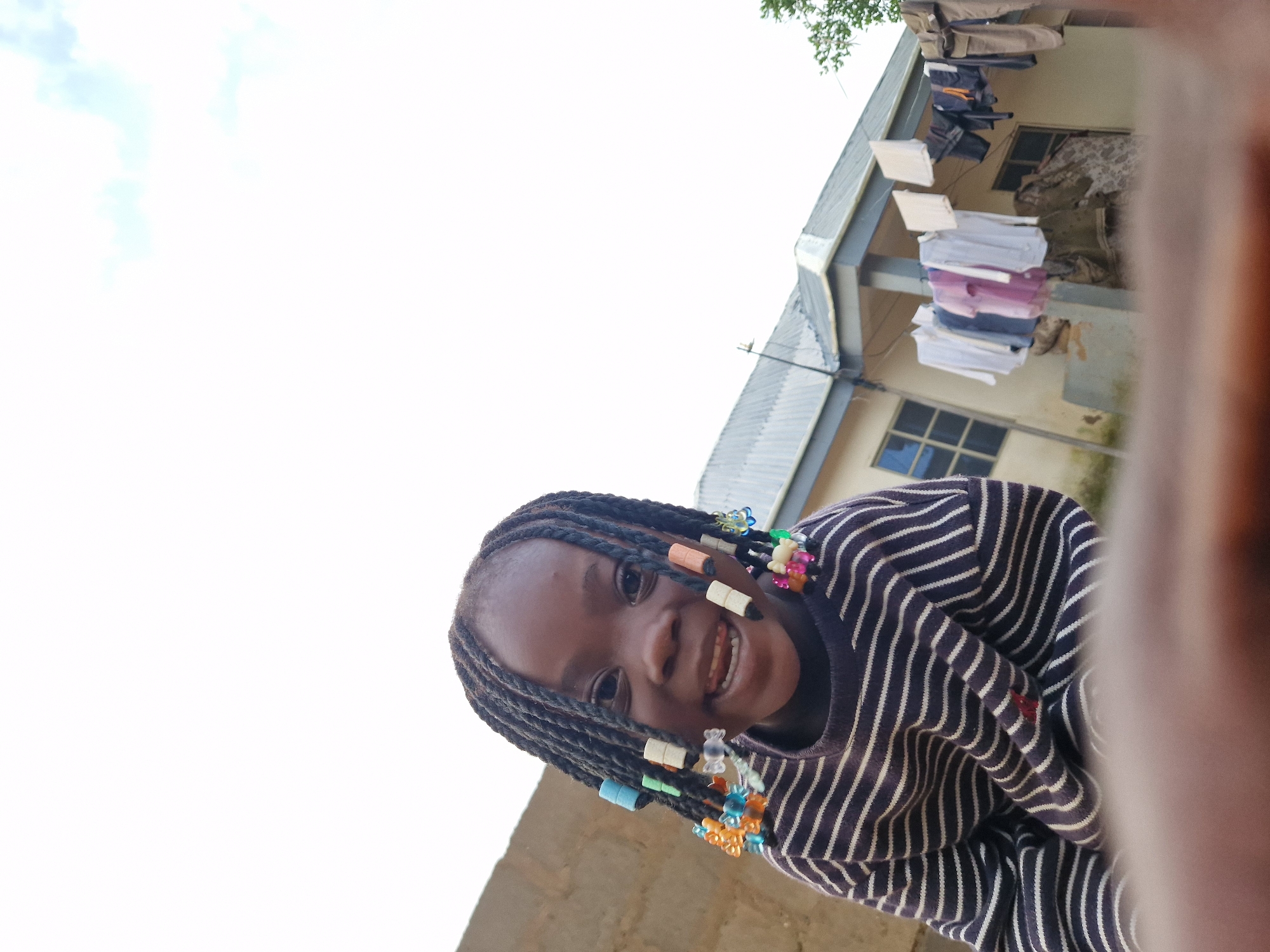 In Jos, Nigeria, a young girl with braided hair adorned with colorful beads is looking at the camera. She is wearing a striped sweater. Behind her, a building with a corrugated metal roof has laundry hanging on a clothesline in front of it. The sky is visible in the background.