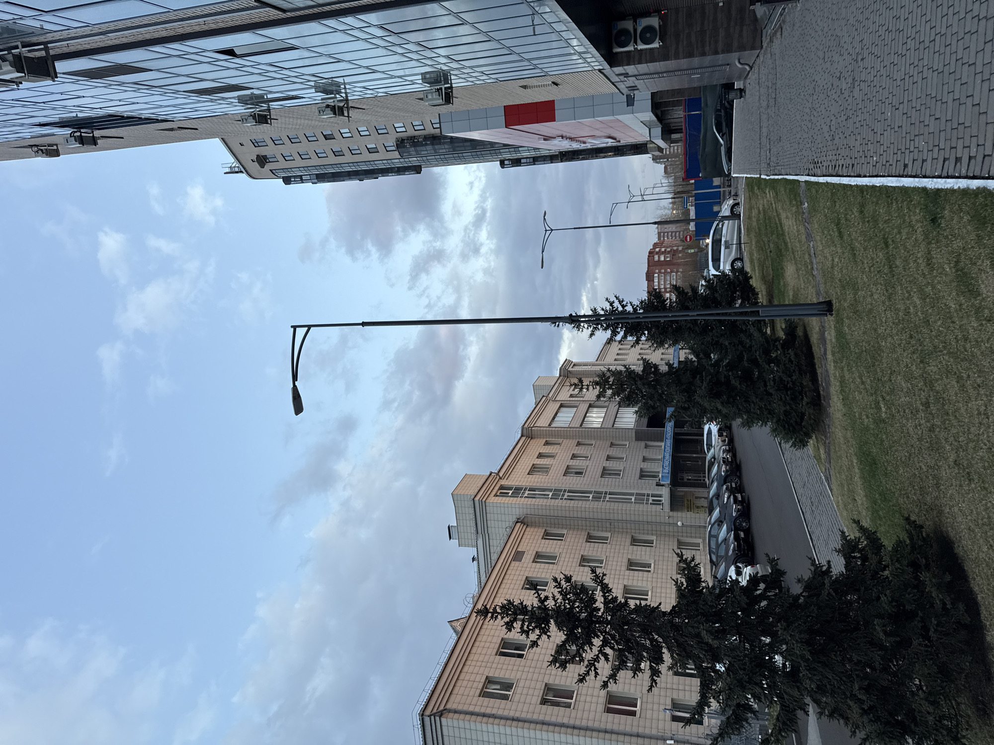 The image shows an urban landscape in Krasnoyarsk, Russia. Two buildings dominate the frame: one with mirrored glass and a red advertisement, and the other an older, tan-colored building with multiple windows. Between the buildings is a street with parked cars and lampposts. Trees are planted along the side of the street. The sky is cloudy, with light blue visible. The image is shot from a low angle and appears to be taken vertically.