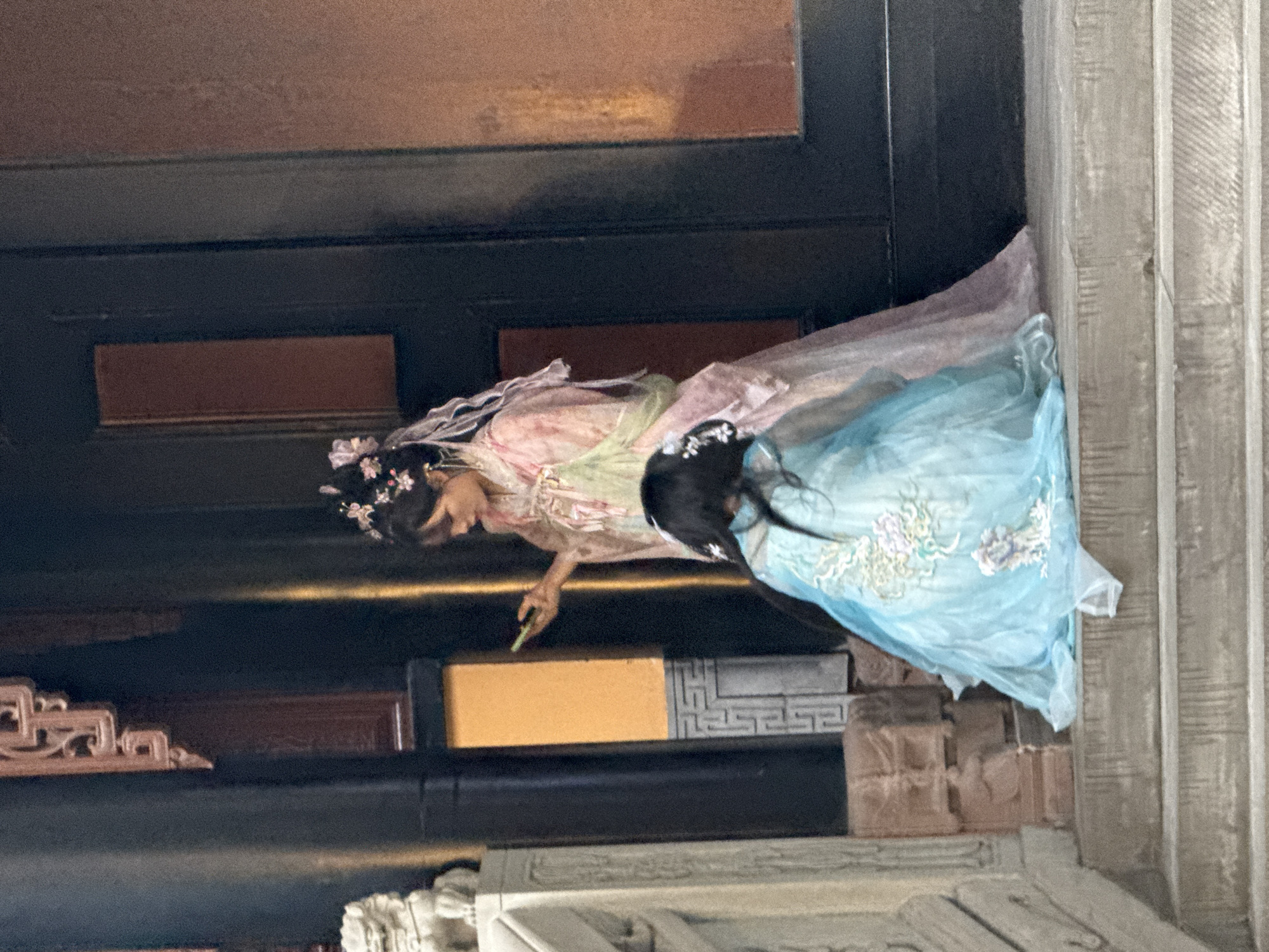 Two individuals, possibly dressed in traditional Chinese attire, are visible on stone steps in front of a dark-colored doorway. The person on the right, standing and facing left, is wearing a pink and green layered robe and has flowers in their hair. They are holding a small, light green object, possibly a mobile phone, in their right hand. The second individual, kneeling on the steps and facing away from the camera, is wearing a flowing light blue dress with floral embroidery. The architectural elements in the background suggest a historical or traditional setting, potentially within Yuzhong District, China, given the stone carvings and the style of the building. The overall lighting suggests an outdoor scene during the day.