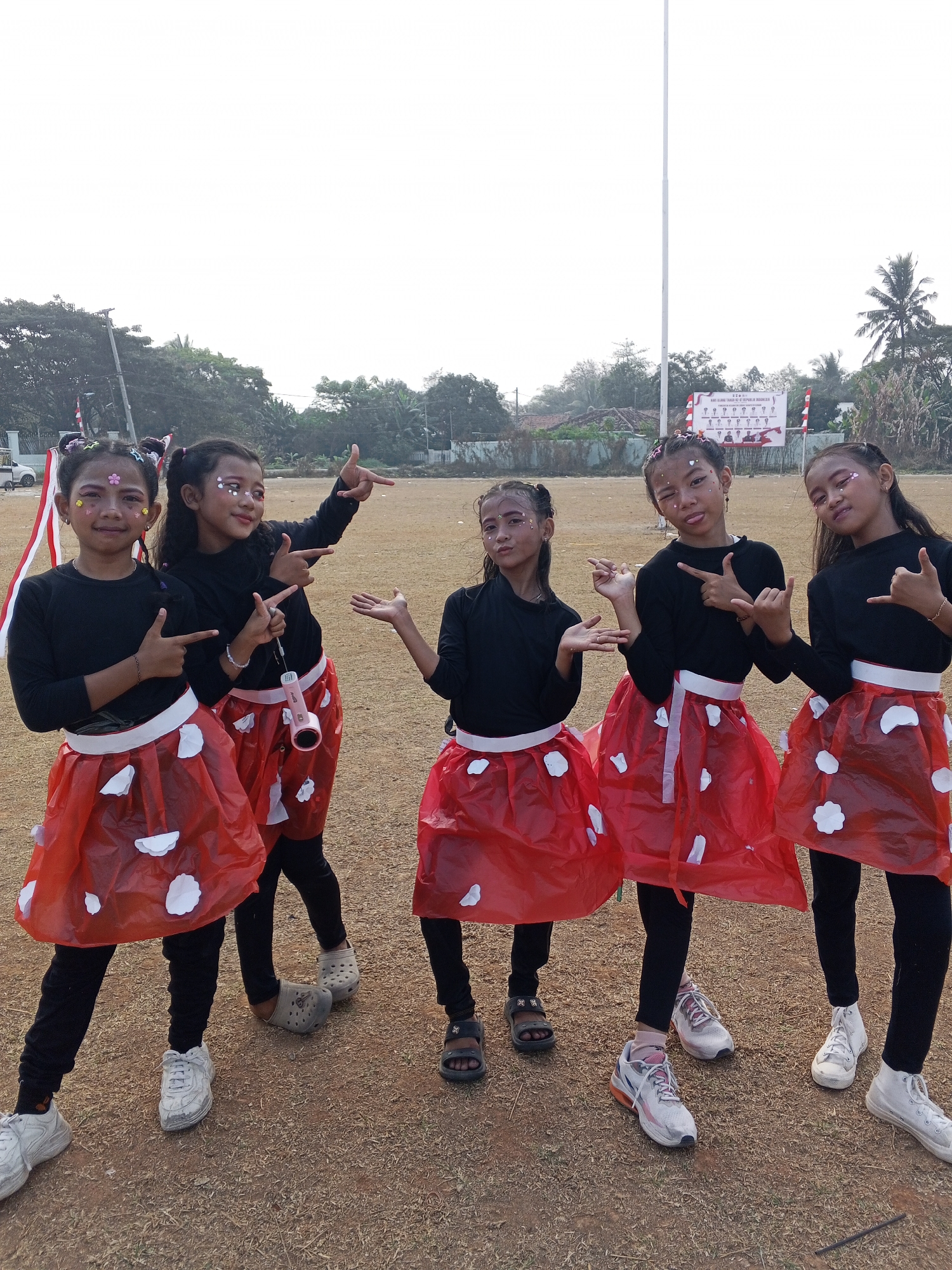 In Ranjeng, Indonesia, five young girls stand in a line outdoors, facing forward and posing for the camera. They are dressed in matching black long-sleeved shirts and bright red skirts adorned with white petal-like cutouts. Each girl has glitter and face paint, with two of them sporting colorful hair accessories. The ground beneath them is covered in dry grass and dirt. In the background, a white flagpole stands tall, accompanied by a banner displaying Indonesian text and small illustrations. Trees and buildings are visible further in the distance.