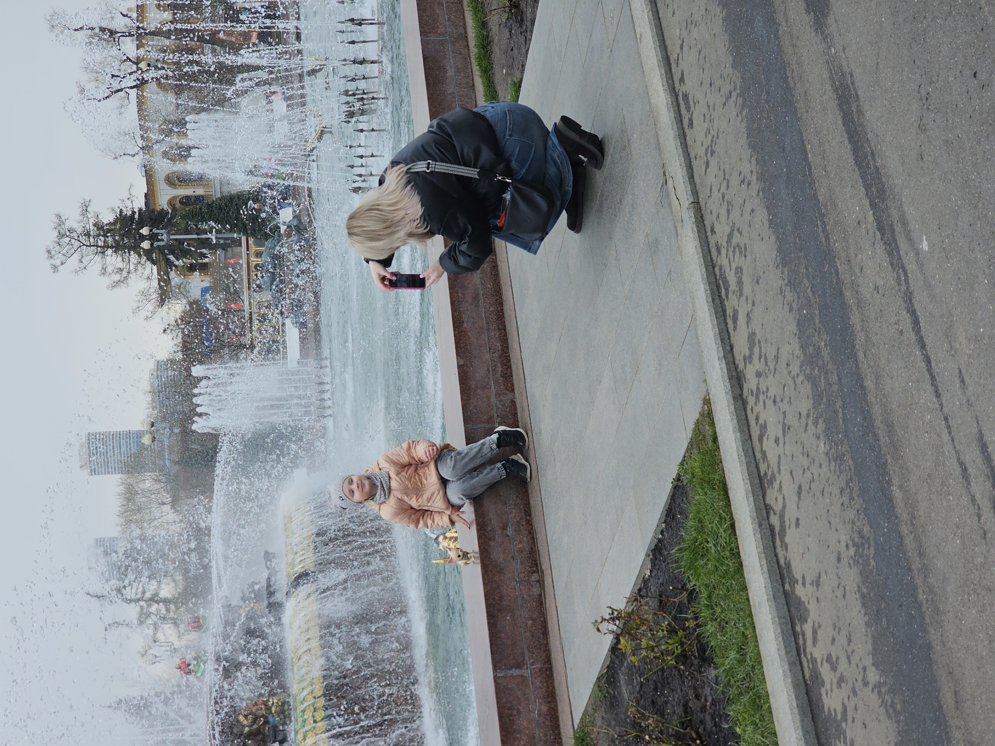 This image captures an outdoor scene from an elevated perspective, showing two individuals by a large, ornate fountain in a city park or square, consistent with a location like Moscow, Russia.
In the foreground, a blonde woman, dressed in a black jacket, blue jeans, and black boots, is kneeling or crouching on a paved pathway. She holds a smartphone with a pink case, seemingly focused on taking a photograph or video of the fountain or the activity beyond it. A black shoulder bag is visible across her body.
To her left, a child is seated on the low wall bordering the fountain. The child is wearing a light brown or peach puffer jacket, grey trousers, black and white sneakers, a grey knit hat, and a grey scarf. A small, light-colored toy, possibly a unicorn, rests beside the child on the fountain's edge. The child is looking towards the camera, appearing calm and engaged with the surroundings.
The central feature of the scene is a large, multi-tiered fountain, vigorously spouting water high into the air, creating a misty atmosphere. The fountain's structure includes decorative elements, with visible gold or yellow patterns along its base. The splashing water partially obscures the background.
Beyond the fountain, several trees, some appearing bare or with minimal foliage, suggest it might be a cooler season like spring or autumn. In the distance, various city buildings, including some high-rise structures, can be seen through the mist and trees. The sky is overcast, resulting in soft, diffused lighting across the scene, indicating daytime without direct sunlight.
The immediate surroundings include a clean, paved sidewalk where the people are, separated by a curb and a narrow strip of green grass from an asphalt road that exhibits some wet patches. There is no discernible text visible in the image. The overall impression is one of leisure and enjoying a public space.