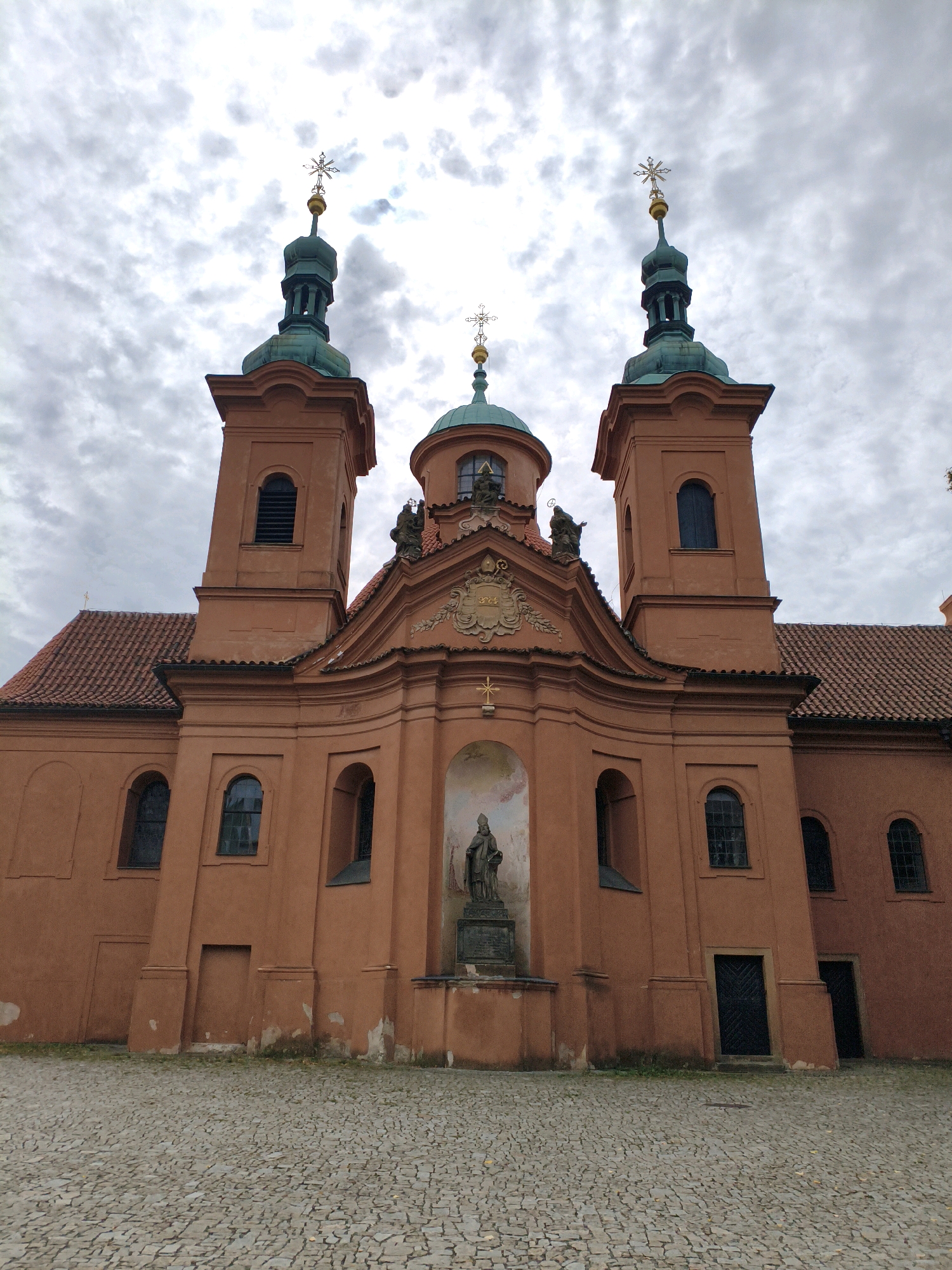 The image shows the Loreta Basilica in Prague, Czechia. The basilica is a terracotta-colored building with two towers topped with green domes and golden crosses. A central dome is also visible. In front of the entrance, there is a statue of a figure standing on a pedestal. The ground in front of the basilica is paved with cobblestones. The sky is overcast with clouds.
