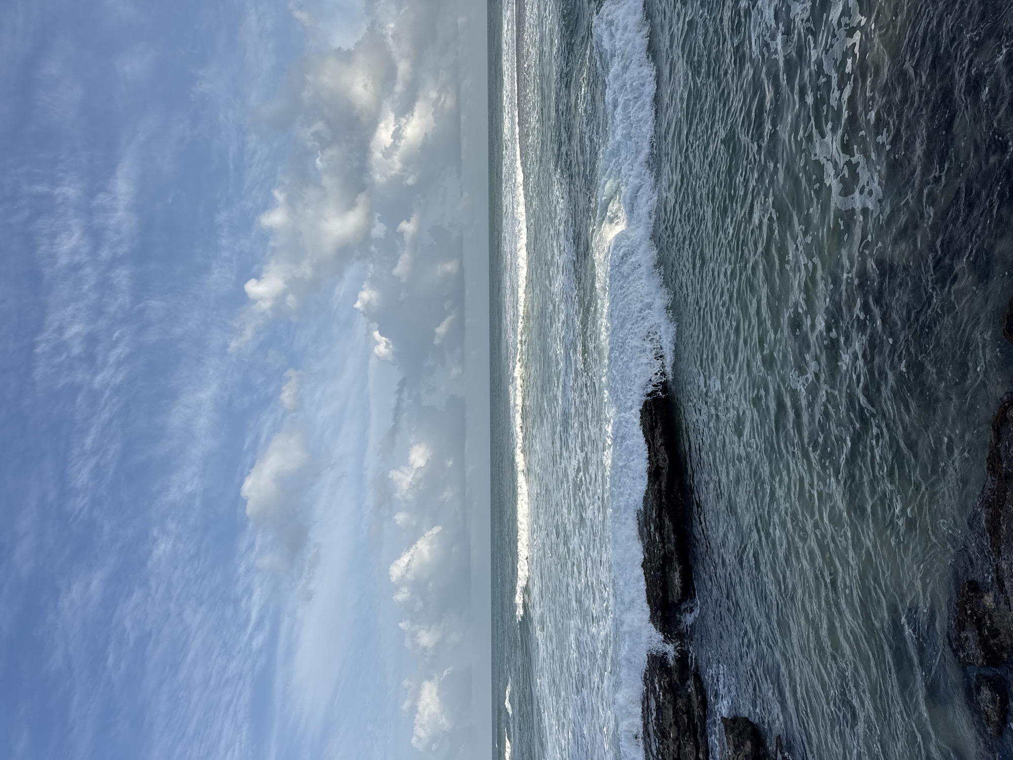 The image depicts a view of the ocean from the shore in Goviyapana, Sri Lanka. Dark, wet rocks are visible in the foreground, partially submerged by the incoming tide. Waves are breaking over these rocks, creating white foam. The water is a deep blue-green, with ripples and foam indicative of movement. In the mid-ground, several lines of waves are rolling towards the shore, some with crests of white foam. The horizon is visible, separating the ocean from the sky. The sky is a pale blue with scattered, wispy white clouds, some of which are tinged with grey, suggesting a partly cloudy day. The overall scene is one of a dynamic coastal environment under a bright sky. There are no people or animals present in the image.