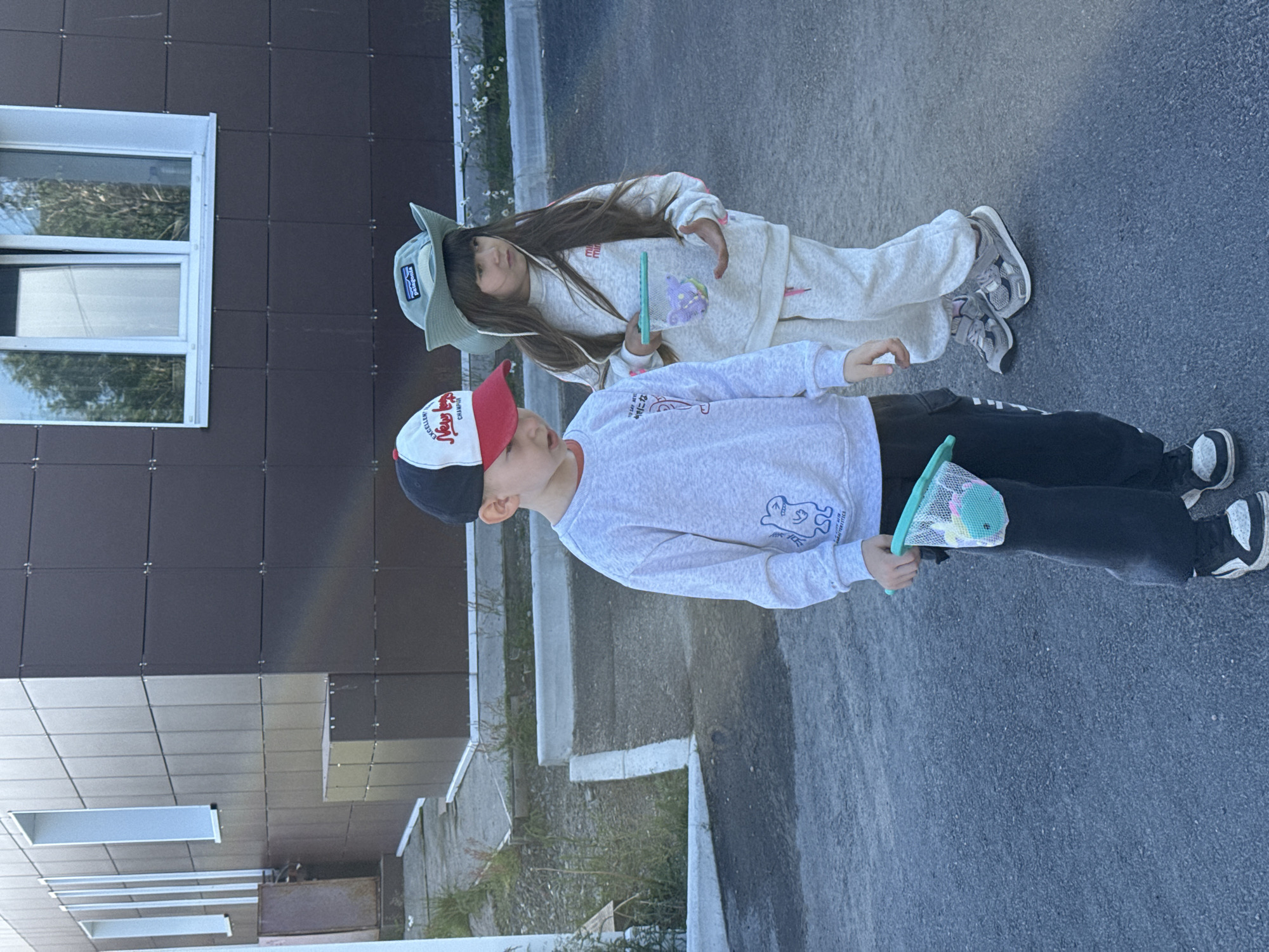 Two children are standing on an asphalt surface in front of a modern building with dark brown tiled walls and white window frames. The child on the left, a boy, wears a gray sweatshirt, black pants, a red and white baseball cap, and black and white sneakers. He holds a small green net with a toy whale inside. The child on the right, a girl, wears a light gray sweat suit, a green wide-brimmed hat with a Patagonia logo, and gray sneakers. She holds a small green net containing a purple toy butterfly. The environment appears to be an urban or suburban setting. There is no specific visual information to definitively place this scene in Teriberka, Russia.