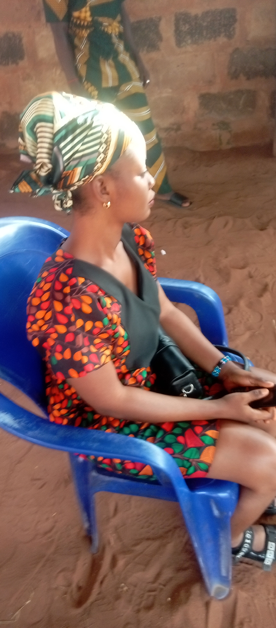 A woman sits in a blue plastic chair outdoors in Nkpor, Nigeria. She is wearing a headwrap, dress with orange, red, and green leafy pattern sleeves, and a black bodice. She has gold earrings and a blue beaded bracelet. A black purse is on her lap. She is looking to the right. The ground is sandy and reddish-brown. Another person is partially visible in the background, wearing a patterned green and yellow outfit. A block wall is also visible in the background.