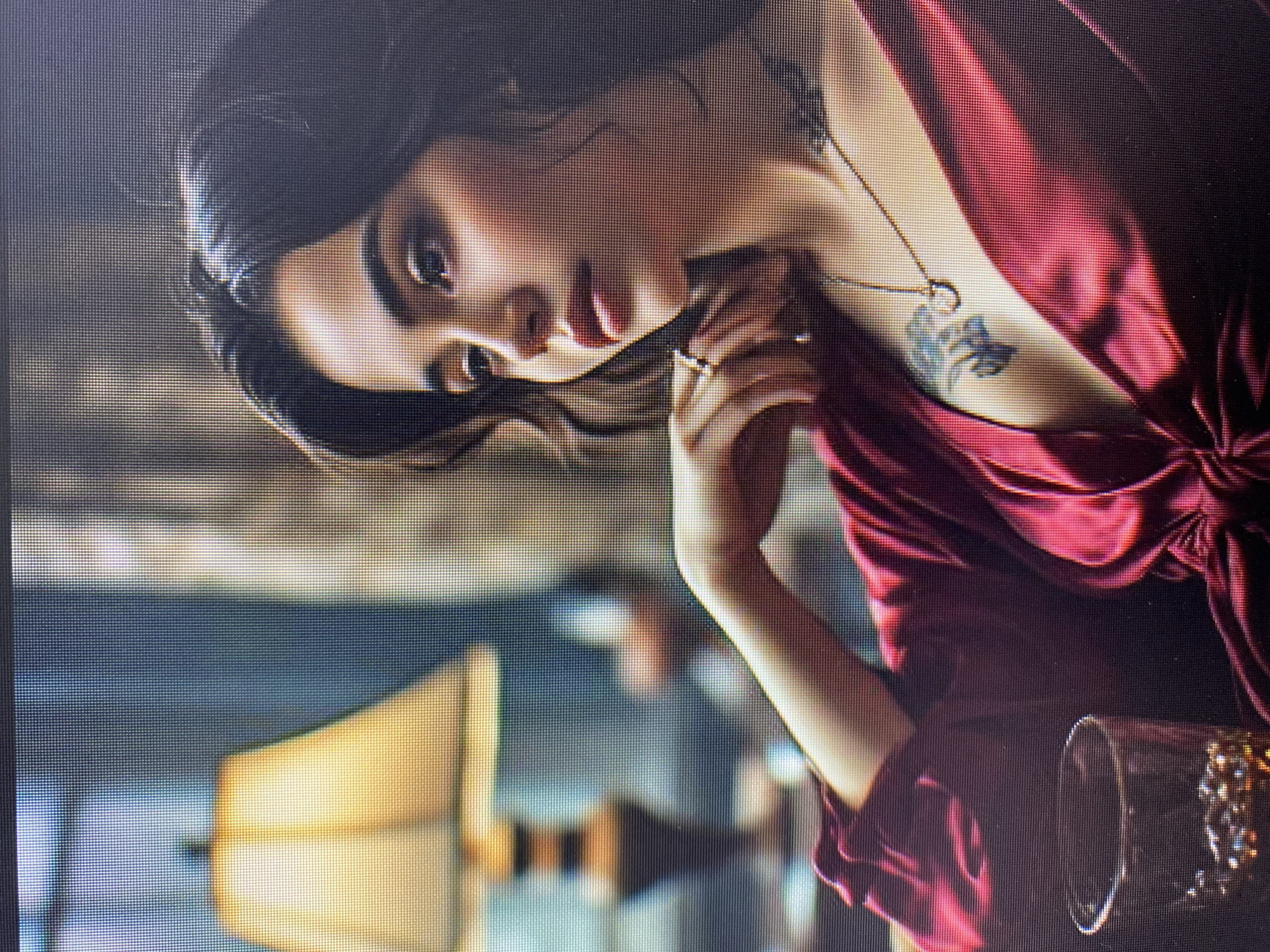 The image shows a close-up of a young woman, posed with her head tilted back and eyes looking towards the viewer. She is wearing a dark red, silky dress with a plunging neckline, revealing a tattoo on her chest and a delicate necklace. Her hand is resting on her chin, and she is wearing a ring on one finger. The lighting is dim, suggesting an indoor setting, possibly at night. The background is blurred, but hints of furniture and possibly a lamp are visible, creating an intimate and somewhat mysterious atmosphere. The woman's expression appears to be alluring or contemplative. The overall aesthetic is elegant and sophisticated.