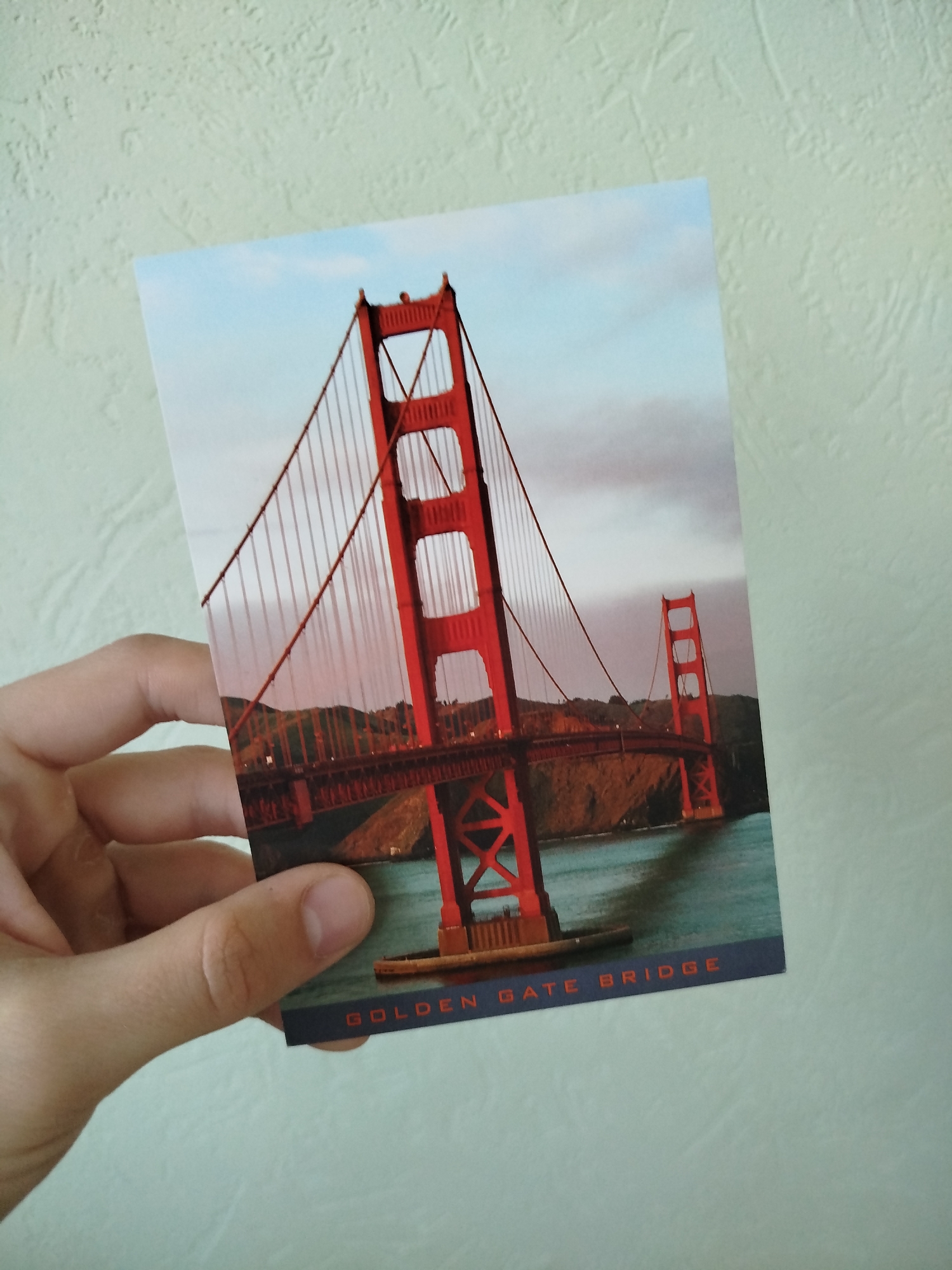 A person's hand holds a postcard depicting the Golden Gate Bridge. The bridge's towers are red, and it spans across a body of water. The postcard is printed with the text "Golden Gate Bridge." The background is a light textured wall.
The scene is not in Varna, Bulgaria. It is in an indoor environment, likely a home or office.
