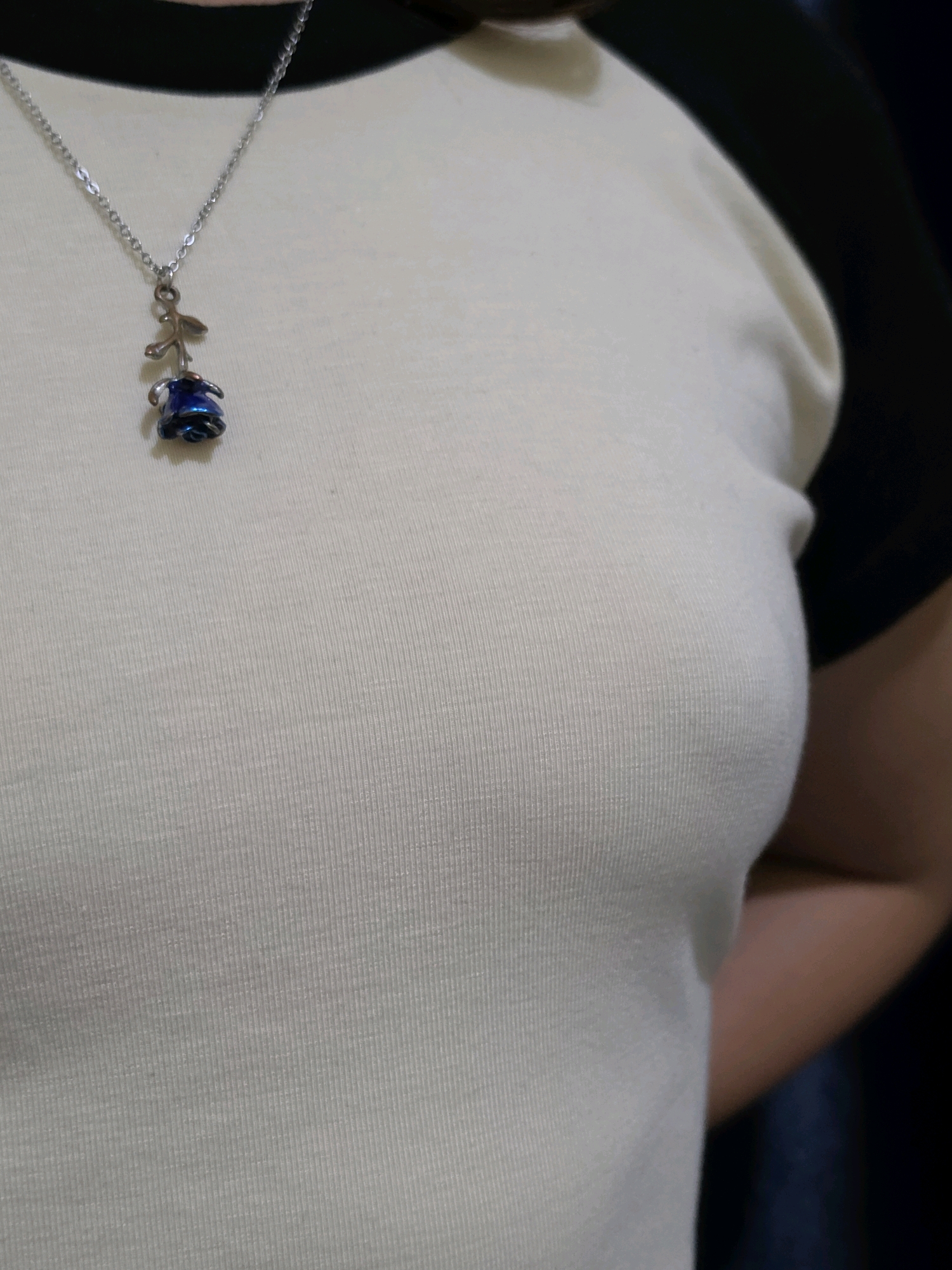This is a medium shot of a person wearing a cream-colored t-shirt with black sleeves. A silver necklace with a small, blue rose pendant hangs around their neck. The background is dark and out of focus. The image is likely taken indoors. There is no information in the image to suggest it is located in Phan Thiết, Vietnam.