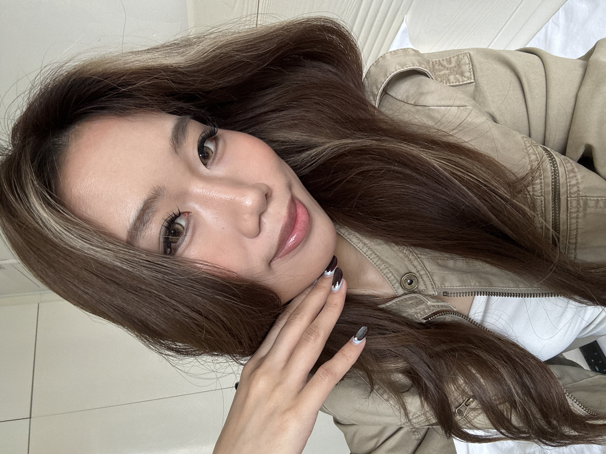 A woman lies down, facing the camera. She has long brown hair with blonde highlights and light skin. She is wearing a beige jacket over a white shirt. Her right hand is resting on her cheek. Her nails are painted with a black and silver design. The background appears to be a white wall, possibly a bedroom. Location: Chenghua District, China cannot be verified from the given image.